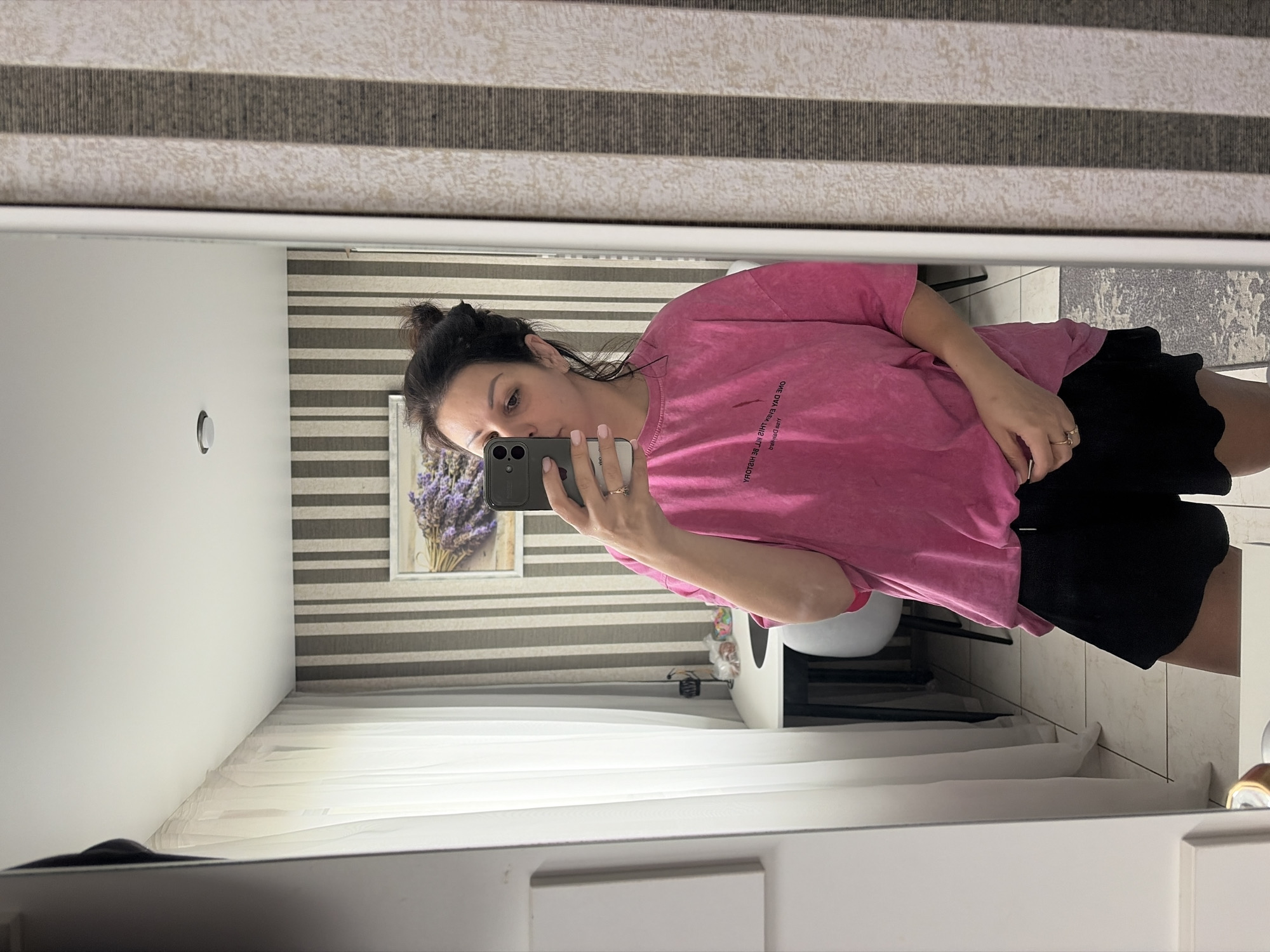 A woman with her dark hair pulled up in a bun takes a mirror selfie in a well-lit room. She is wearing a pink oversized t-shirt with text and black shorts. She is holding a smartphone in her right hand to take the picture. The background includes a wall with vertical striped wallpaper, sheer white curtains, and a framed picture of lavender. The floor is tiled. A table and chairs are visible in the reflection of the mirror. The overall impression is one of a casual indoor setting, possibly a dressing room or bedroom.