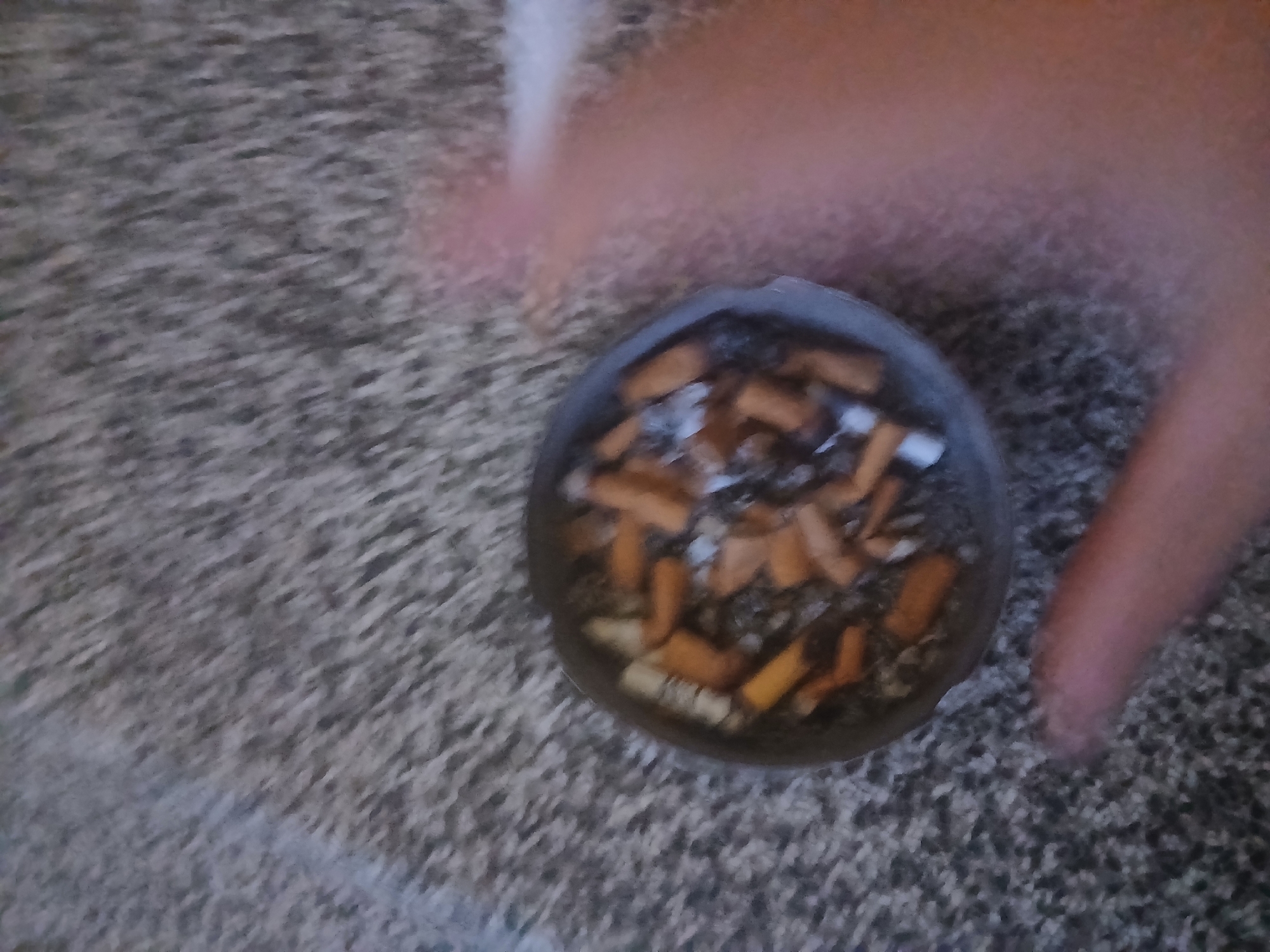 The image shows an ashtray filled with cigarette butts, resting on a textured gray carpet. A blurred hand is visible in the right side of the frame. The ashtray is dark gray and circular. The cigarette butts vary in color, with some showing white filters and others appearing brown and orange from use. Ash is also present in the ashtray. The carpet has a shaggy, speckled appearance. The overall scene suggests a casual indoor environment. There is no information to place this image in Pilsen, Czechia.
