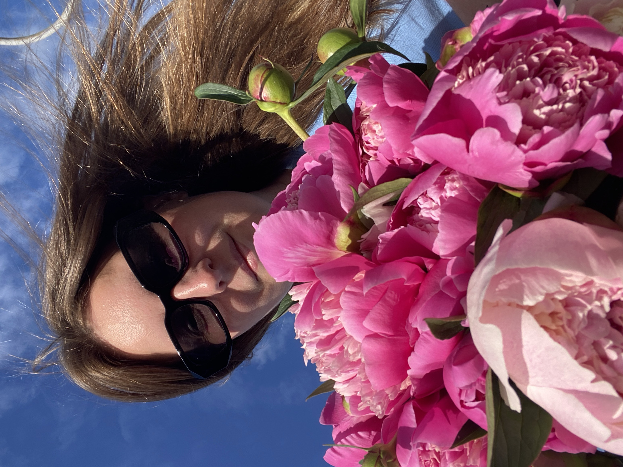 A close-up, low-angle shot captures a woman holding a bouquet of bright pink peonies against a bright blue sky with some wispy white clouds. The woman, seen from the shoulders up, wears black sunglasses and has long, brown hair blowing in the wind. Her face is partially obscured by the flowers and her sunglasses. The peonies are the dominant subject, with many fully bloomed flowers in various shades of pink, and a few closed buds visible. The background is a clear, sunny sky. There are no discernible landmarks indicating a specific location within Noviy, Russia.