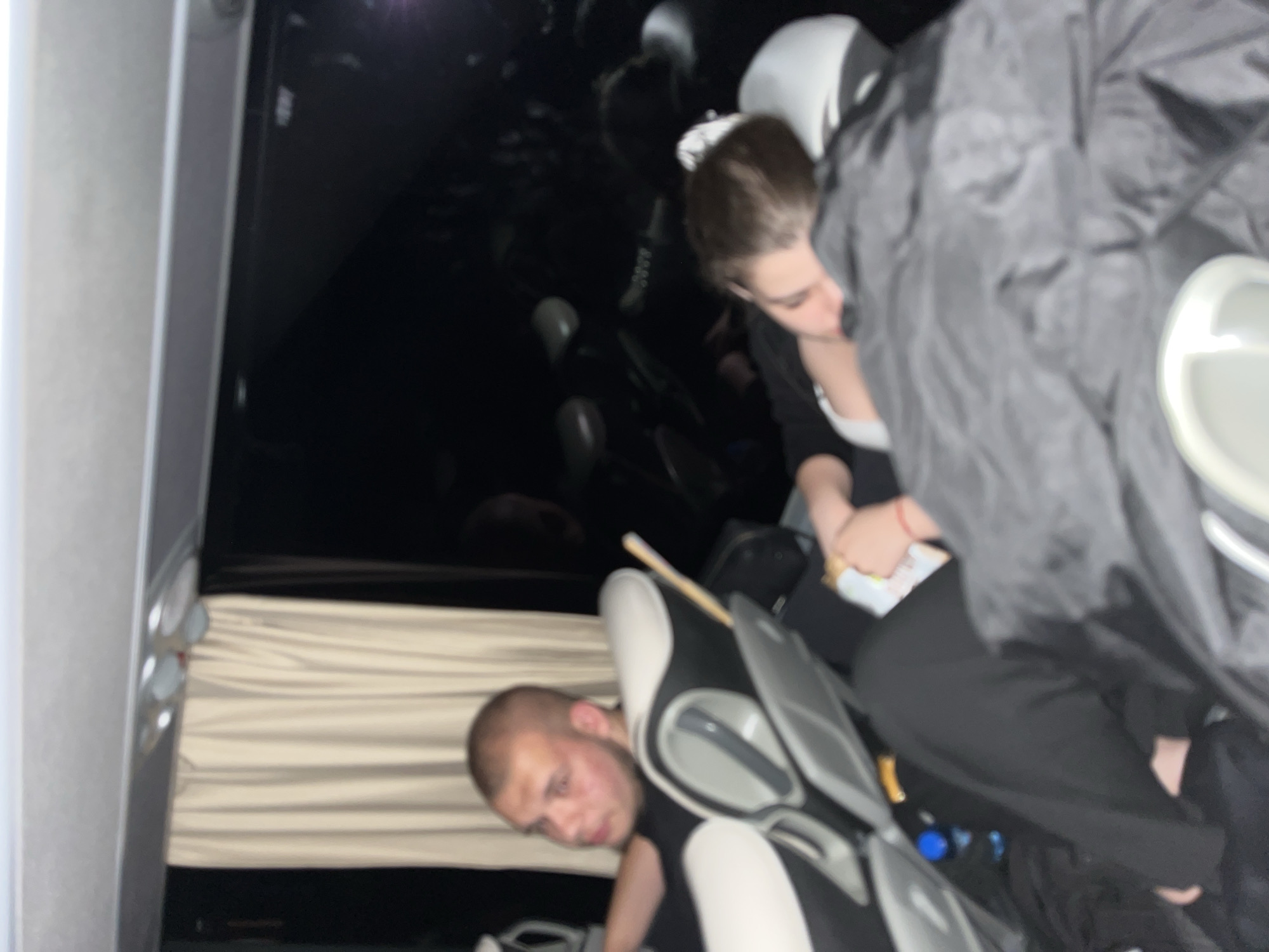 The image shows the interior of a bus. A man with a shaved head looks towards the camera from behind a seat. A woman is seated, appearing to hold something in her hands. Seats can be seen down the length of the bus. The lighting appears dim. There is no confirmed evidence to establish the location as Popovitsa, Bulgaria.