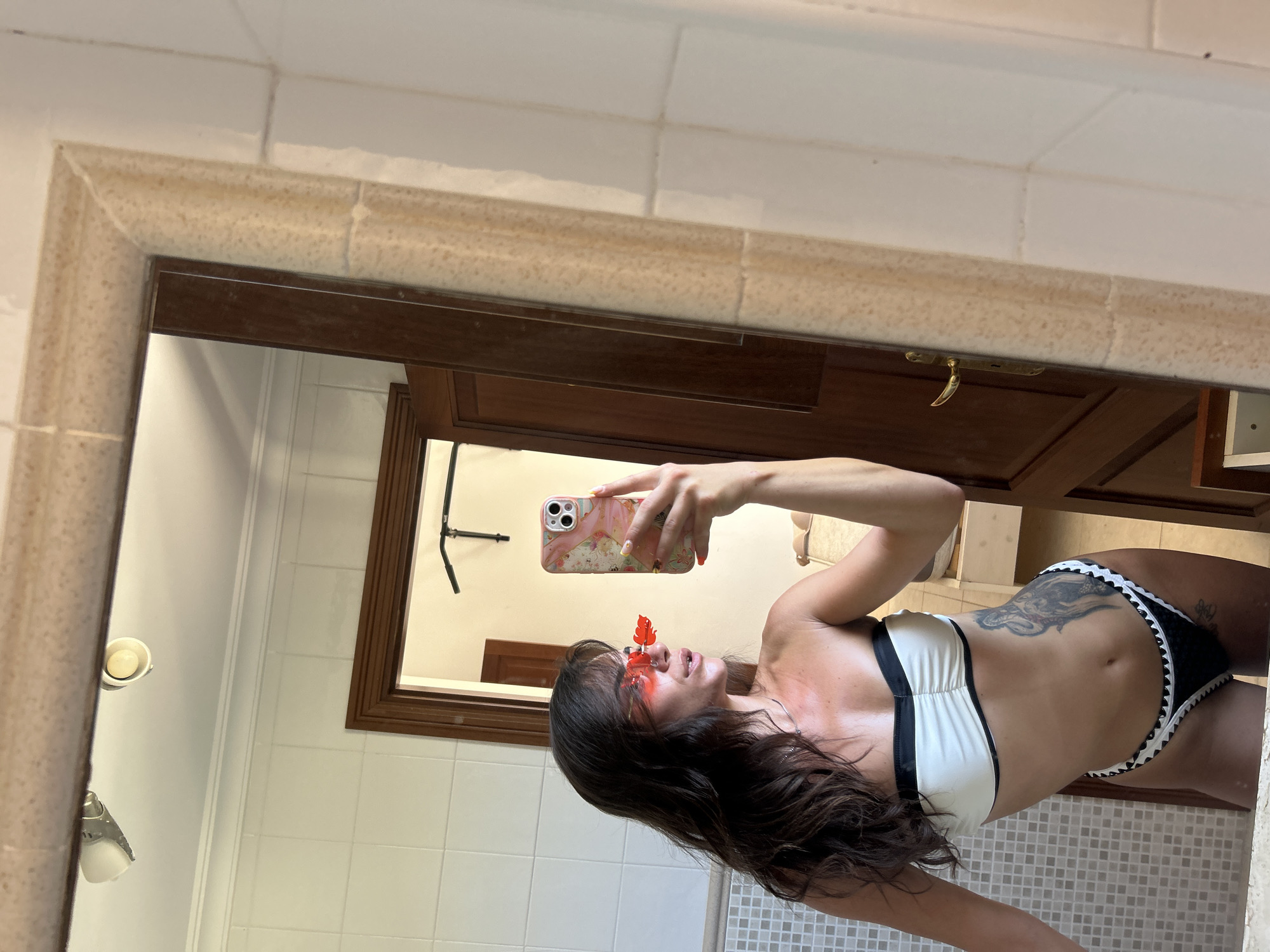 A young woman with long, dark hair and sunglasses takes a mirror selfie in what appears to be a bathroom. She is wearing a black and white bikini with ruffled trim. Her left arm is raised, holding a phone with a floral case. A tattoo is visible on her left side. The bathroom features white tiled walls, a wooden door with a brass handle, and a large mirror. The scene is illuminated by overhead lights. The location is identified as Capdepera, Spain.