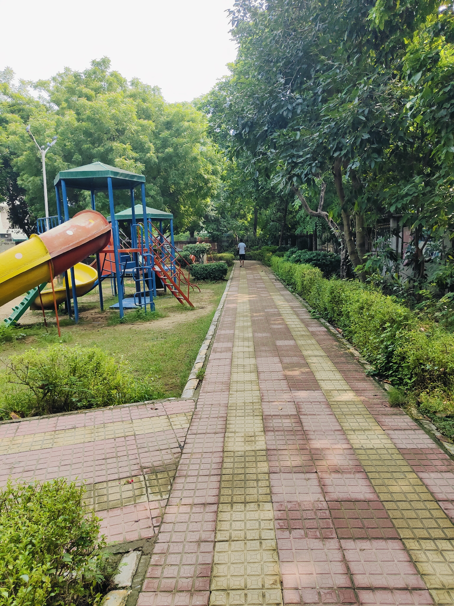 The image depicts a paved walkway in a park or residential area in Gurgaon, India. The walkway is constructed of interlocking square tiles in alternating patterns of reddish-brown and light yellow. To the left of the walkway, there is a children's playground with a yellow and red slide, blue metal structures, and green steps. Lush green trees and bushes line the right side of the walkway, with a fence visible further in the background. The sky is overcast. In the distance, a lone individual wearing a white t-shirt and dark shorts is walking away from the camera down the pathway. The scene suggests a tranquil setting, possibly early morning or late afternoon.