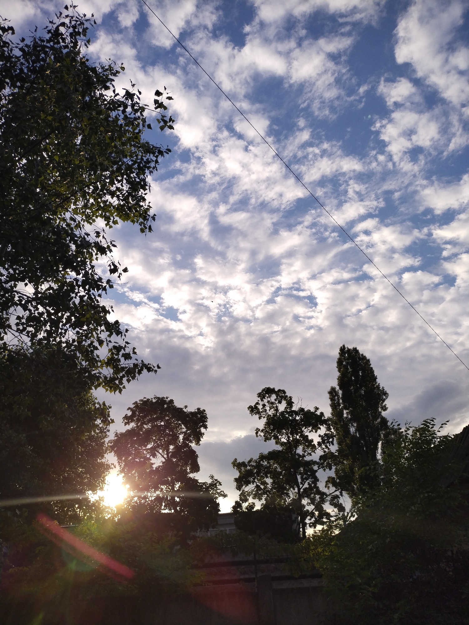The image depicts a sky filled with a mixture of blue sky and scattered white clouds, with a single black wire stretching diagonally across it. In the foreground, silhouetted against the bright sky, are the tops of several trees. The sun is visible, peeking through the leaves of a large tree on the left side of the frame, creating a lens flare effect with streaks of light. The lower portion of the image is dominated by the dark silhouettes of trees and foliage, with hints of a building or structure in the background, possibly a fence or wall. The scene suggests an outdoor setting, likely a park or residential area in Horishni Plavni, Ukraine, during daylight hours, possibly late afternoon or early morning given the angle of the sun. No people are visible in the image.