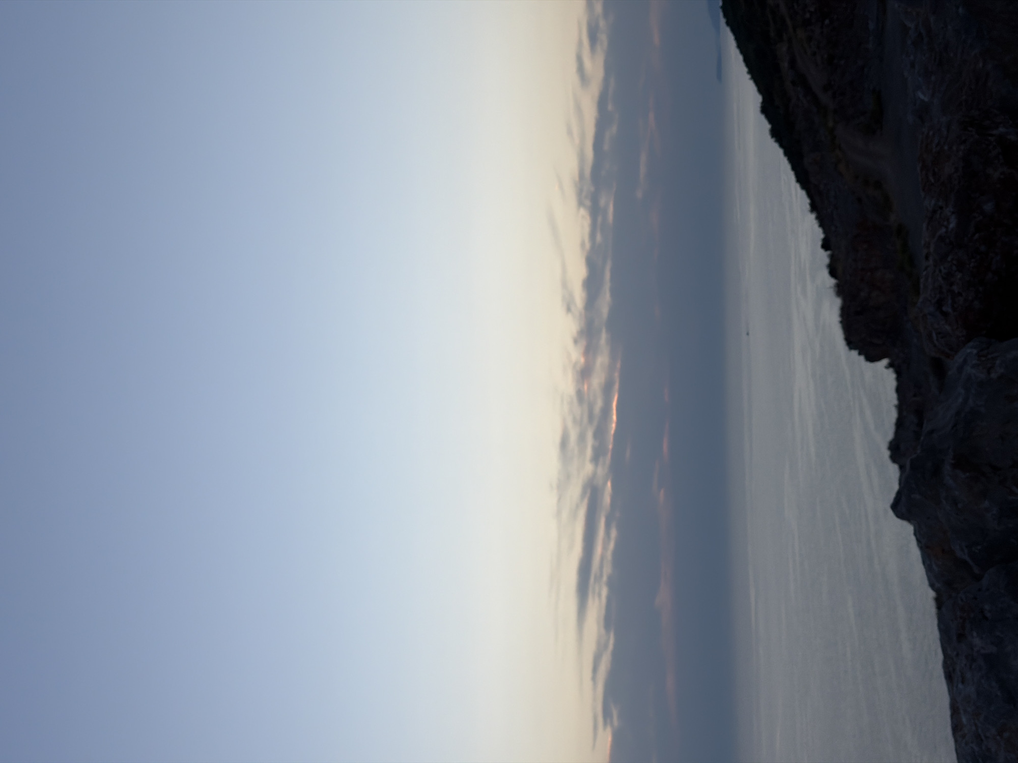 The image depicts a serene coastal scene at dusk. The sky transitions from a pale blue at the top to a soft orange and pink hue near the horizon, where a thin layer of clouds is visible. The calm sea stretches out to meet the sky, with gentle ripples reflecting the fading light. In the foreground, rugged rocks form a natural embankment overlooking the water. To the right, a sloping hillside covered in sparse vegetation descends towards the sea. The overall atmosphere is tranquil, suggesting a peaceful evening in a coastal location. Based on the prompt's request to include the location, this scene is situated in the city of Zagora, Montenegro.