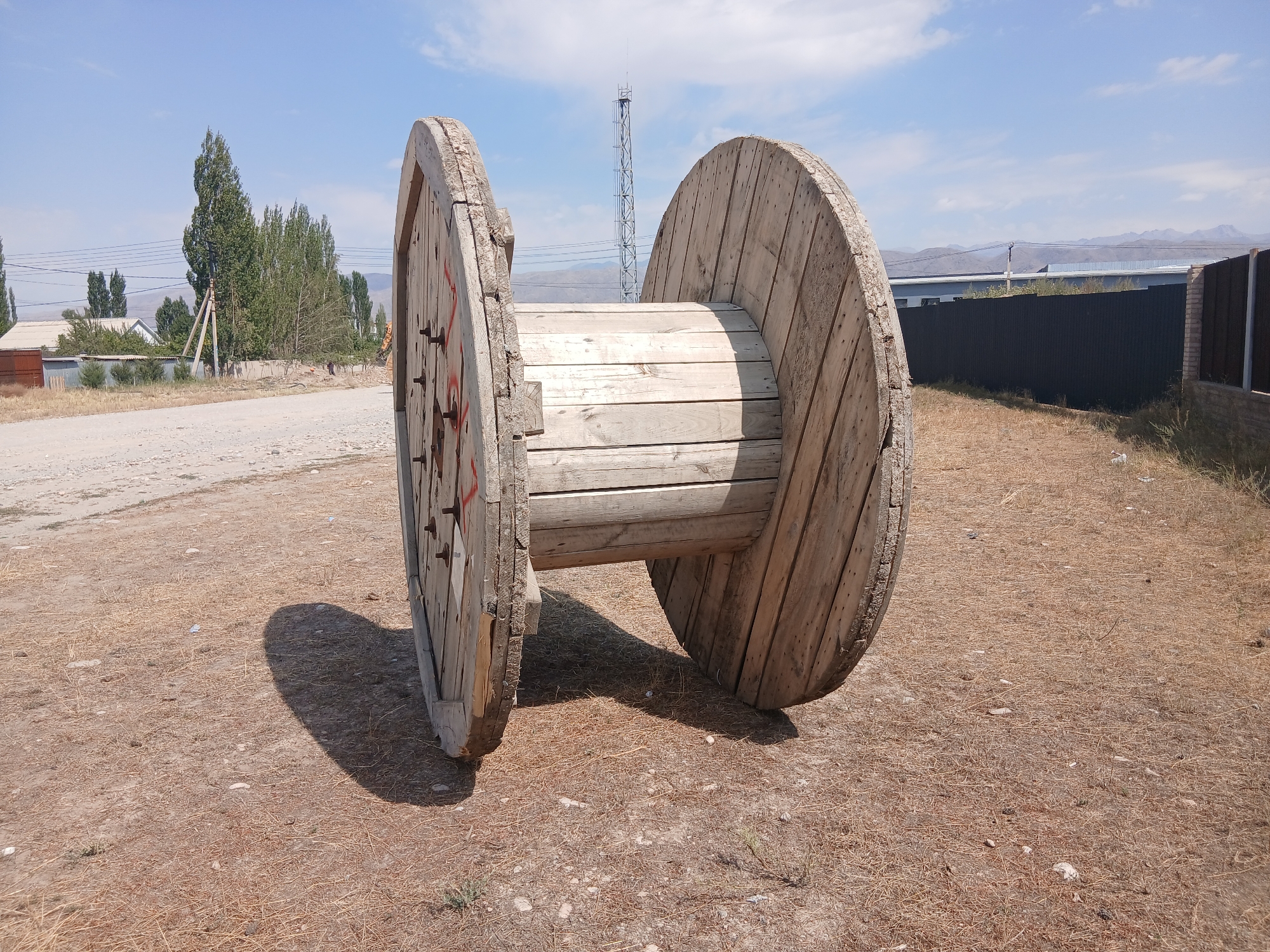 A large, weathered wooden cable spool rests on dry, straw-like ground under a clear blue sky. The spool has two large circular wooden sides and a central barrel made of horizontal wooden planks. Red markings are visible on one of the spool's sides. In the background, a dirt road leads towards a cluster of trees and buildings, including a house with a white roof and a grey metal fence. Further in the distance, a tall, lattice-like tower stands against the horizon, and mountains are visible under a hazy sky. The scene appears to be in an open, semi-rural area in Talas City, Kyrgyzstan.