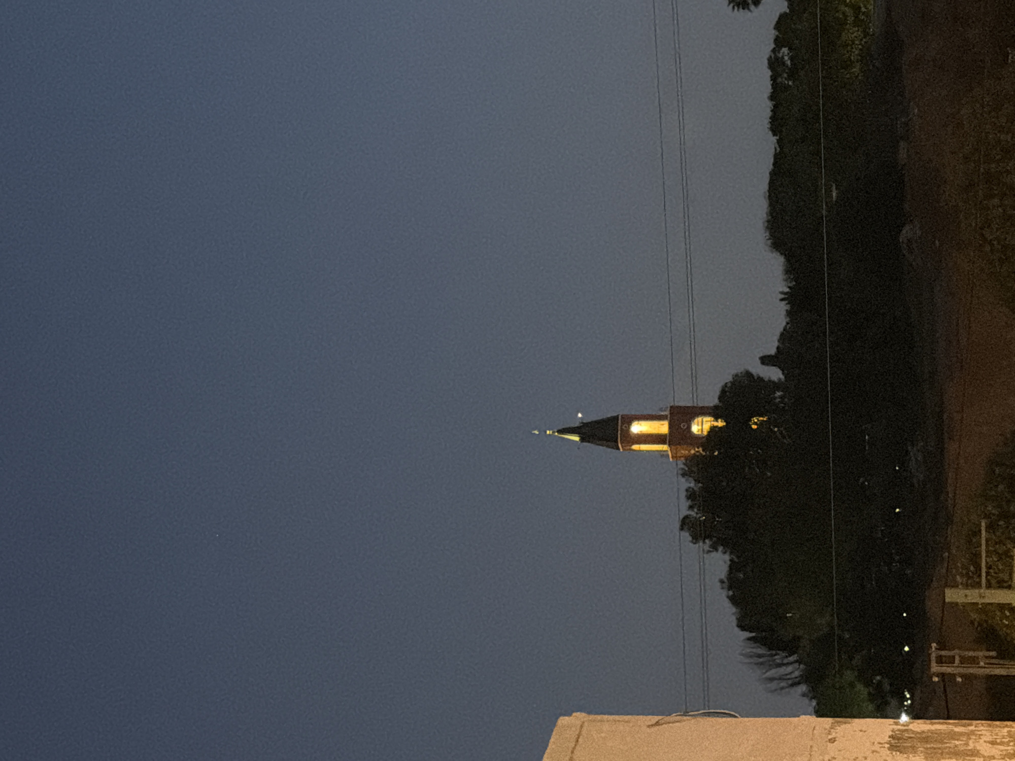 The image depicts a tranquil evening or nighttime scene, primarily composed of a dark blue sky and a prominent, illuminated tower. The sky occupies the majority of the left and upper frame, transitioning from a dark blue at the top to a slightly lighter shade near the horizon line.
A tall, slender tower, possibly a clock tower or minaret, is the central subject, positioned on the right side of the frame. Its upper section is dark with a pointed, spired roof. The windows of the tower glow with a warm, golden yellow light, suggesting internal illumination. The lower part of the tower is partially hidden behind dense, dark green foliage, which appears to be a cluster of trees or bushes, filling the mid-right section of the image. Faint, scattered light points can be discerned within this dark foliage, possibly distant lights or reflections.
Several thin, dark lines, resembling power lines, stretch across the right side of the frame, running vertically against the sky and above the treeline. In the bottom foreground, a light-colored building structure, likely a wall or rooftop, is partially visible, grounding the scene.
The time of day is clearly evening or night, indicated by the dark sky and the artificial lighting of the tower. The weather appears clear, with no clouds visible. There is no discernible activity or event taking place; the scene is static and peaceful. No text is visible in the image. Given the context, this serene urban landscape is likely located in Tel-Aviv, Israel.