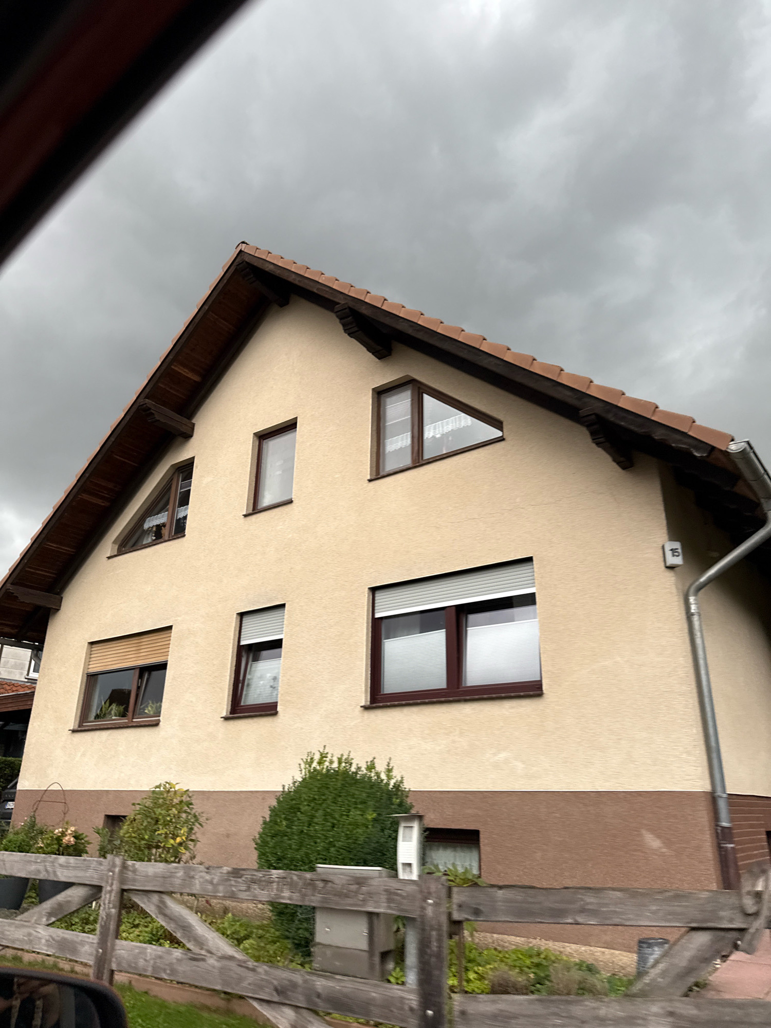 The image captures a beige, two-story house in Bad Sachsa, Germany, under a cloudy sky. The house has a dark brown roof with visible wooden eaves. There are several windows on the upper floor, including a triangular window on the left gable. The lower portion of the house is painted a contrasting brown. In front of the house, a rustic wooden fence runs horizontally across the lower part of the frame, with green shrubs and some potted plants behind it. A metal downspout runs vertically down the side of the house, next to a house number that appears to be "15". The overall scene suggests a residential area on an overcast day.