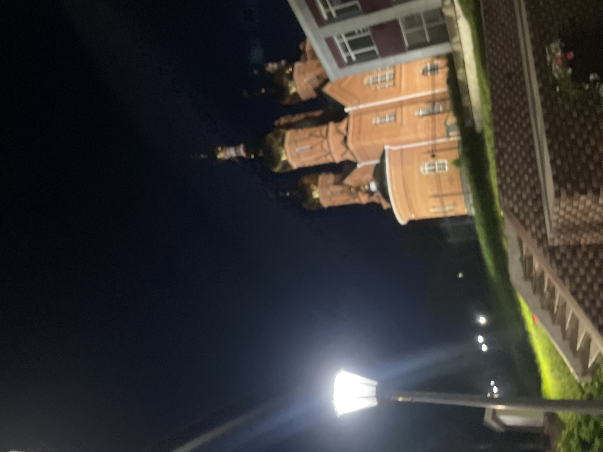 The image depicts a nighttime view of the Church of the Resurrection in Krasnye Baki, Russia. A brightly lit street lamp is in the foreground on the left, casting a strong white light. Behind the lamp, a red brick church with golden domes is visible. To the right of the church, a modern building with white-framed windows and red accents is partially visible. In the middle ground, a grassy slope leads up to the church, and a set of concrete stairs is visible on the right side of the slope. The sky is dark blue, indicating it is night. There are no people visible in the image.