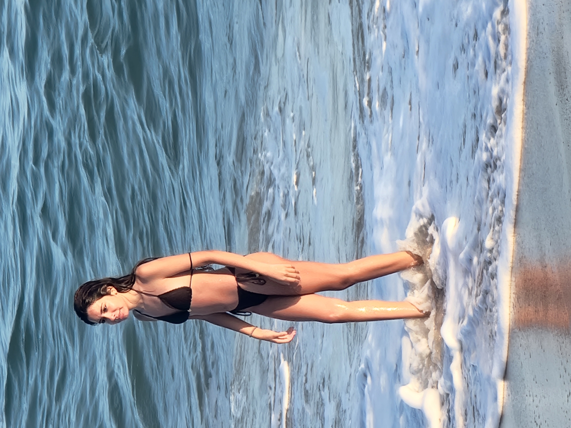 A young woman with dark hair and a black bikini stands in the shallow water at the edge of the ocean. She is facing to the left, looking out at the water. The water is blue and choppy, with white foam washing over her legs and feet. The sand is wet and reflects the light. The image appears to have been taken on a beach in Quarteira, Portugal, given the characteristic wavy patterns of the Atlantic Ocean.