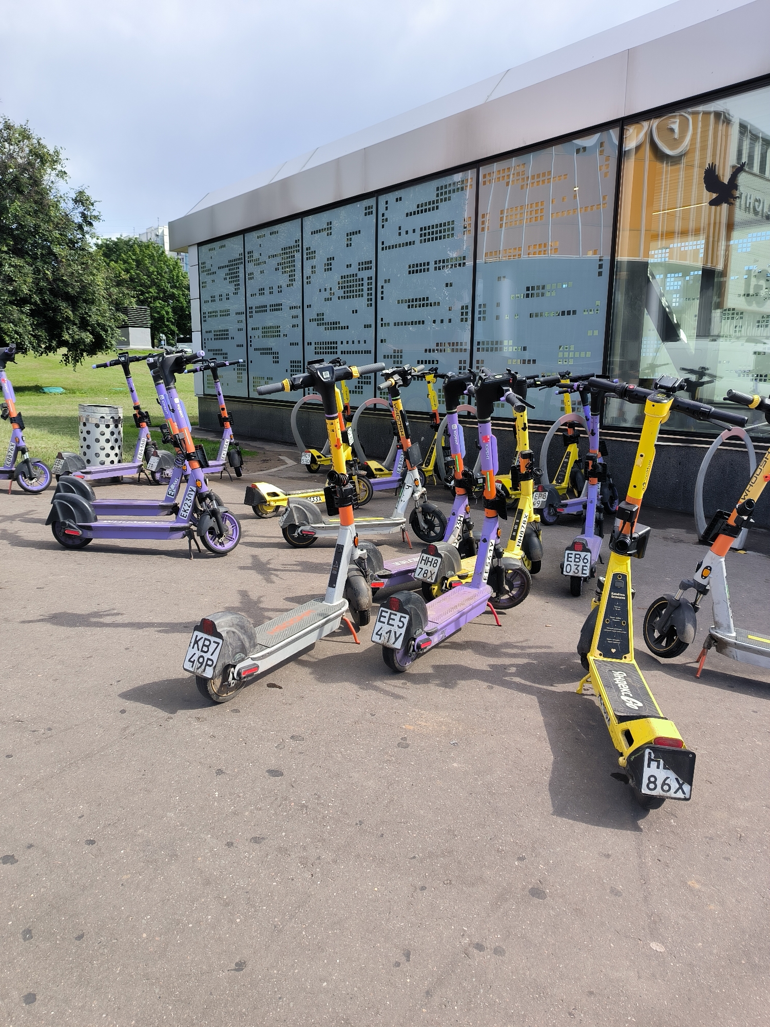 This image shows a row of electric scooters parked on a paved surface, likely a sidewalk or plaza, in Moscow, Russia. The scooters are a mix of purple and yellow, with some brands like "Whoosh" and "Юрент" (YooRent) visible on them. Several of the scooters have license plates with alphanumeric codes such as "KB7 49P", "EE5 41Y", "EB6 03E", and "H 86X". In the background, there is a modern building with a glass facade that reflects the sky and surrounding environment, including a stylized graphic of a bird. Trees and a trash can are also visible, suggesting an outdoor urban setting. The lighting indicates it is daytime, possibly late morning or afternoon, under a bright, partly cloudy sky. No people are visible in the frame, and the overall scene conveys a sense of urban mobility and rental services. The environment appears clean and orderly.