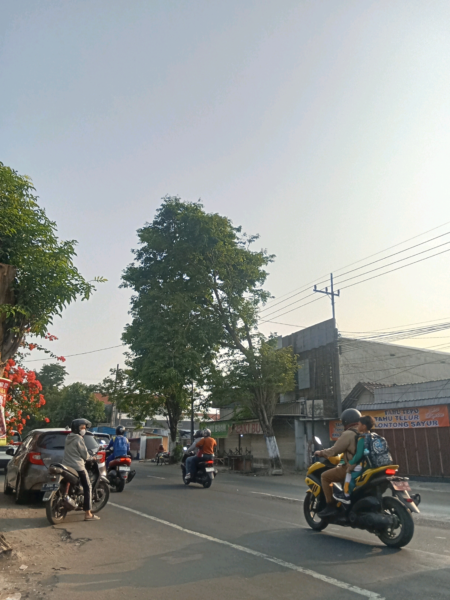 The image captures a street scene in Kaliwungu, Indonesia, bathed in the soft light of what appears to be early morning or late afternoon. The sky is a pale, hazy blue, indicating a potentially warm day.
In the foreground, a paved road stretches into the distance, with a distinct white line marking the lanes. To the left, a grey car is parked on the side of the road. A person on a black motorcycle, wearing a grey hoodie, black pants, and a white mask, is seated on the motorcycle, facing away from the camera. Another motorcycle with a rider in a blue shirt and helmet is to the left of the car, also facing away. Further down the road, another person on a red motorcycle rides away from the camera, wearing an orange shirt and helmet.
To the right side of the road, a yellow motorcycle is in motion, carrying two people. The rider, wearing a brown jacket and helmet, is in front, while a child with a backpack is seated behind. The motorcycle has a black license plate.
The background features several buildings, including a long, grey structure with a weathered facade and a sign that reads "TAHU TEPO TAHU TELUR LONTONG SAYUR." There are also utility poles with multiple wires running across the scene. Lush green trees are visible on both sides of the street, with a large tree dominating the center of the frame. To the far left, bright red flowers are in bloom on a tree or bush. The overall impression is a busy urban street with moderate traffic and everyday life unfolding.