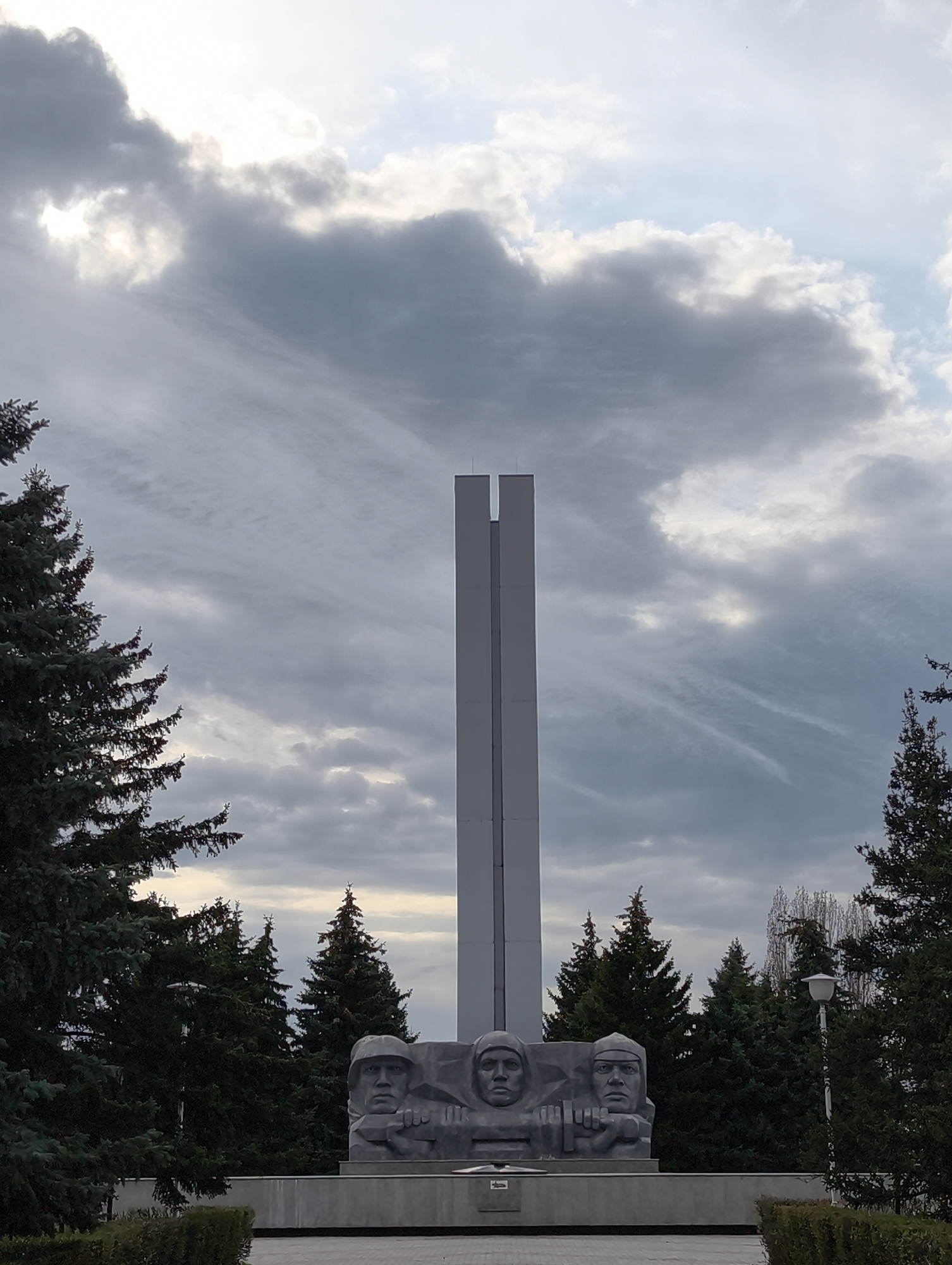 The image shows a monument in Balashov, Russia. The monument consists of three sculpted heads of soldiers set against a long, grey, rectangular tower in a park-like setting. Coniferous trees flank both sides of the monument, with an overcast sky above. The base of the monument is concrete, and a lamppost stands to the right. The overall lighting suggests a cloudy or overcast day.