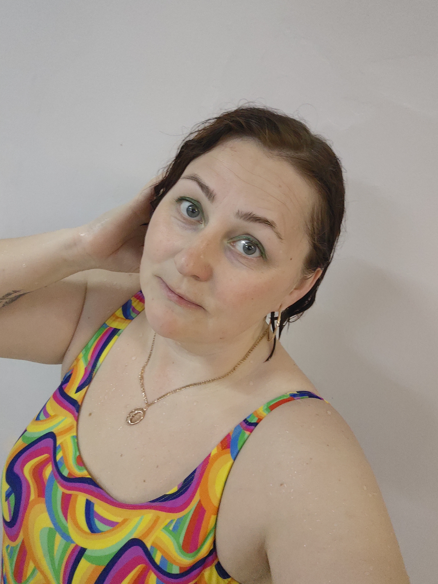 A medium shot depicts a woman with wet, short brown hair wearing a colorful, swirling-patterned swimsuit and gold jewelry, including earrings and a necklace. She is in front of a plain white wall and appears to be in a bathroom or shower. Her right hand is raised, fingers touching her wet hair. Water droplets are visible on her skin and hair. The woman is looking at the camera with a neutral expression, and her eyes appear to have green eye shadow. The image was taken in Markovskii, Russia.