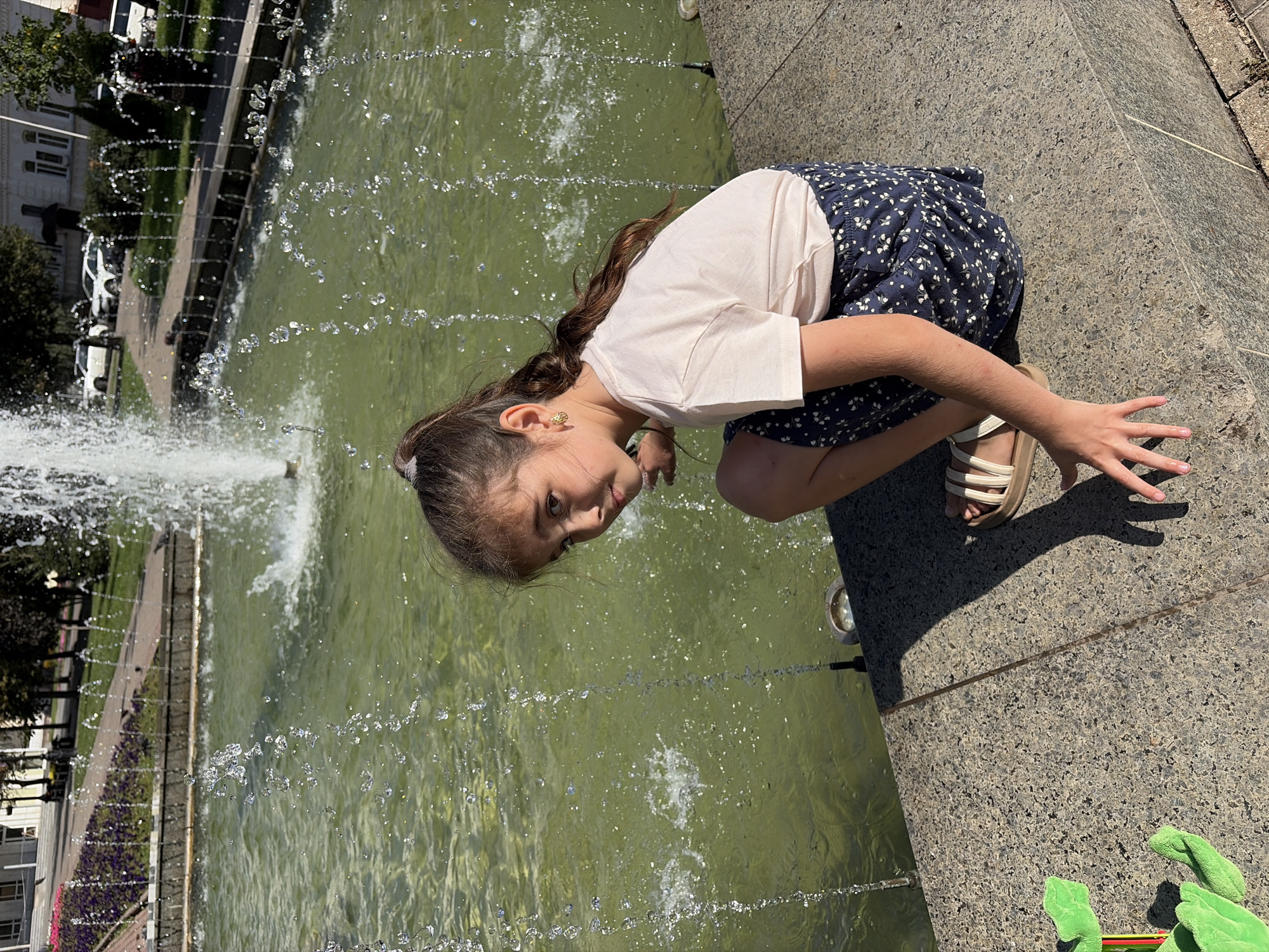 A young girl with long brown hair is crouched at the edge of a fountain, leaning over to observe the water. She is wearing a light-colored t-shirt and a dark blue skirt with a white floral pattern. On her feet are beige sandals. Her right hand is extended towards the water, with her fingers spread. The fountain has multiple streams of water arcing and splashing into a greenish pool. In the background, a park-like setting is visible with trees, a walkway, and some buildings. The lighting suggests it is daytime, likely sunny given the shadows. The girl appears curious and engaged with the water. A bright green stuffed toy is partially visible in the lower right corner of the frame. The image was taken in Tambov, Russia.