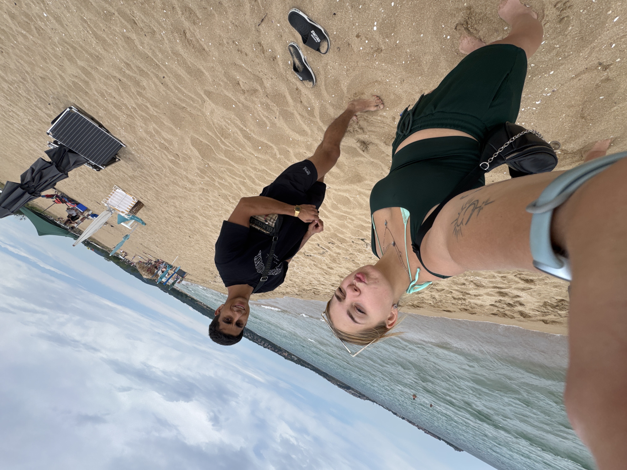The image captures a beach scene in Sozopol, Bulgaria. Two individuals, a man and a woman, are posing for a selfie on the sandy shore. The man, wearing a black t-shirt and black shorts, is smiling and looking towards the camera. The woman, in a green bikini and dark green shorts, is making a kissing face. In the background, the sea stretches out, with gentle waves lapping the shore. Further back, a coastline with vegetation is visible, and on the beach, several beach chairs and umbrellas are set up, indicating a popular tourist spot. A pair of black flip-flops lie discarded on the sand near the couple. The sky is partly cloudy, with a mix of blue sky and white clouds. The overall atmosphere suggests a casual day at the beach.