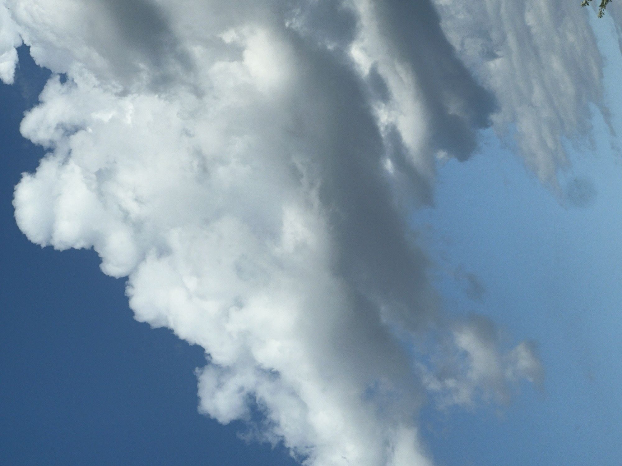 The image shows a sky filled with a variety of clouds. The sky is a bright blue, with large, fluffy white cumulus clouds dominating the upper portion of the frame. Towards the bottom and right side of the image, there are darker, more gray clouds, suggesting potential for rain or a change in weather. The perspective is looking upwards, as if from ground level, capturing a significant portion of the sky. No specific landmarks or identifiable features of Warsaw, Poland are visible in this image.