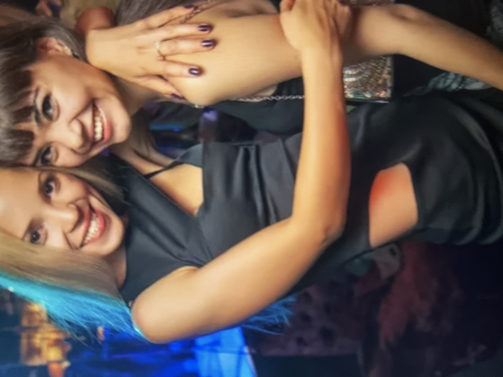 Two young women are posing together, with one woman's arm around the other's shoulder. They are both smiling and appear to be in a dimly lit, possibly urban, environment. The woman on the left has blonde hair and is wearing a black dress with a cut-out detail. The woman on the right has dark hair and is wearing a black top. Their arms are intertwined, and one woman is wearing a ring on her left hand. The background is out of focus and appears to be a public place, potentially a bar or club, with blurred lights and furniture. There is no indication of the location being in Qarağandı city administration, Kazakhstan.