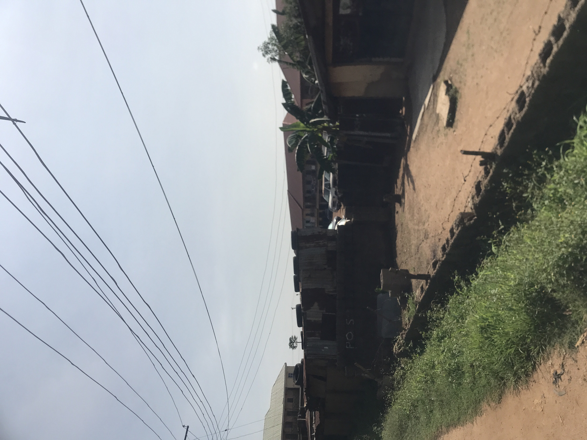 The image depicts an outdoor scene in Rafin Par, Nigeria, characterized by a dirt road on the left, overgrown green grass, and a low-lying wall with barbed wire. Beyond the wall, a compound area is visible, featuring a building constructed with corrugated metal and what appears to be discarded tires. The word "POST" is spray-painted on the corrugated metal structure. Several vehicles, including SUVs, are parked in the background. Further back, multiple-story buildings with red roofs are present, alongside a solitary tree in the distance. Overhead, a network of power lines stretches across the sky, contributing to the urban or semi-urban environment. The lighting suggests it is daytime, with a bright, hazy sky.