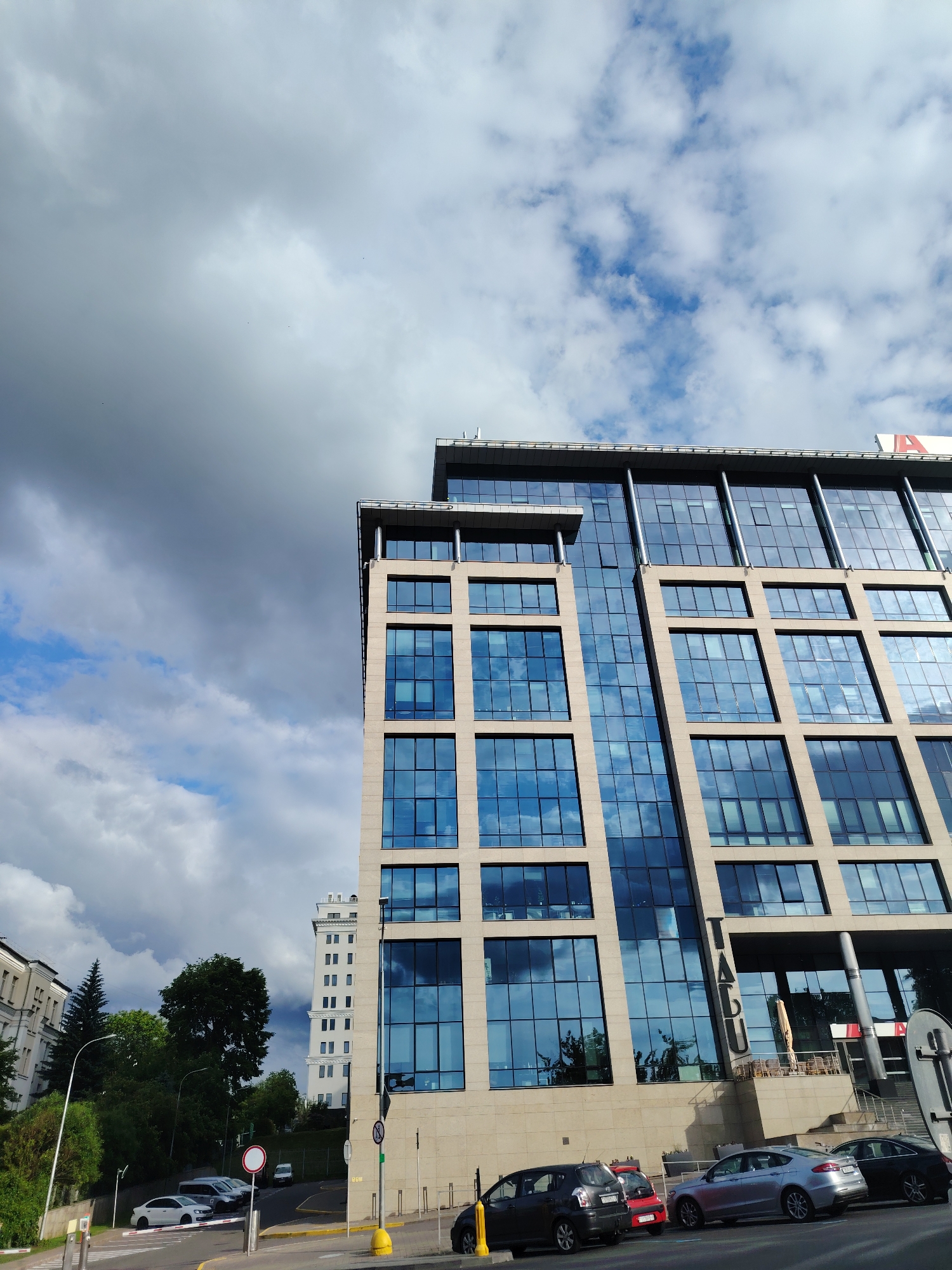 This image captures a modern multi-story office building, identified as the IACU Business Center, located at Pobediteley Avenue, 100, in Minsk, Belarus. The building occupies the right and central portions of the frame, featuring a facade composed of beige stone panels and numerous blue-tinted glass windows that reflect a dynamic sky. Vertically oriented letters spelling "IACU" are prominently affixed to the building's lower-mid facade. A partial red "A" is visible on a sign at the building's rooftop level on the upper right.
The sky above is a significant element, displaying a stark contrast between patches of bright blue with scattered white cumulus clouds on the right and dense, dark grey storm clouds on the left. Small, dark specks, possibly birds, are visible against the clouds in the upper left.
At street level, several cars are parked alongside the building, including a dark grey hatchback, a red hatchback, a silver sedan (Ford Fusion/Mondeo), and a dark sedan. Yellow curb markings define the parking area. A white barrier gate, a yellow bollard, and various street signs, including a red circular sign, are visible in the foreground.
To the left of the main building, the landscape slopes upwards, densely covered with green trees and shrubs. Beyond these, two older-style buildings are partially visible: a light-colored, multi-story building with classical architectural details and, further back, a taller, slender white building with numerous windows. A street light pole is situated among the trees on the left. No specific individuals are discernible performing actions or interactions within the scene. The lighting indicates daylight conditions.