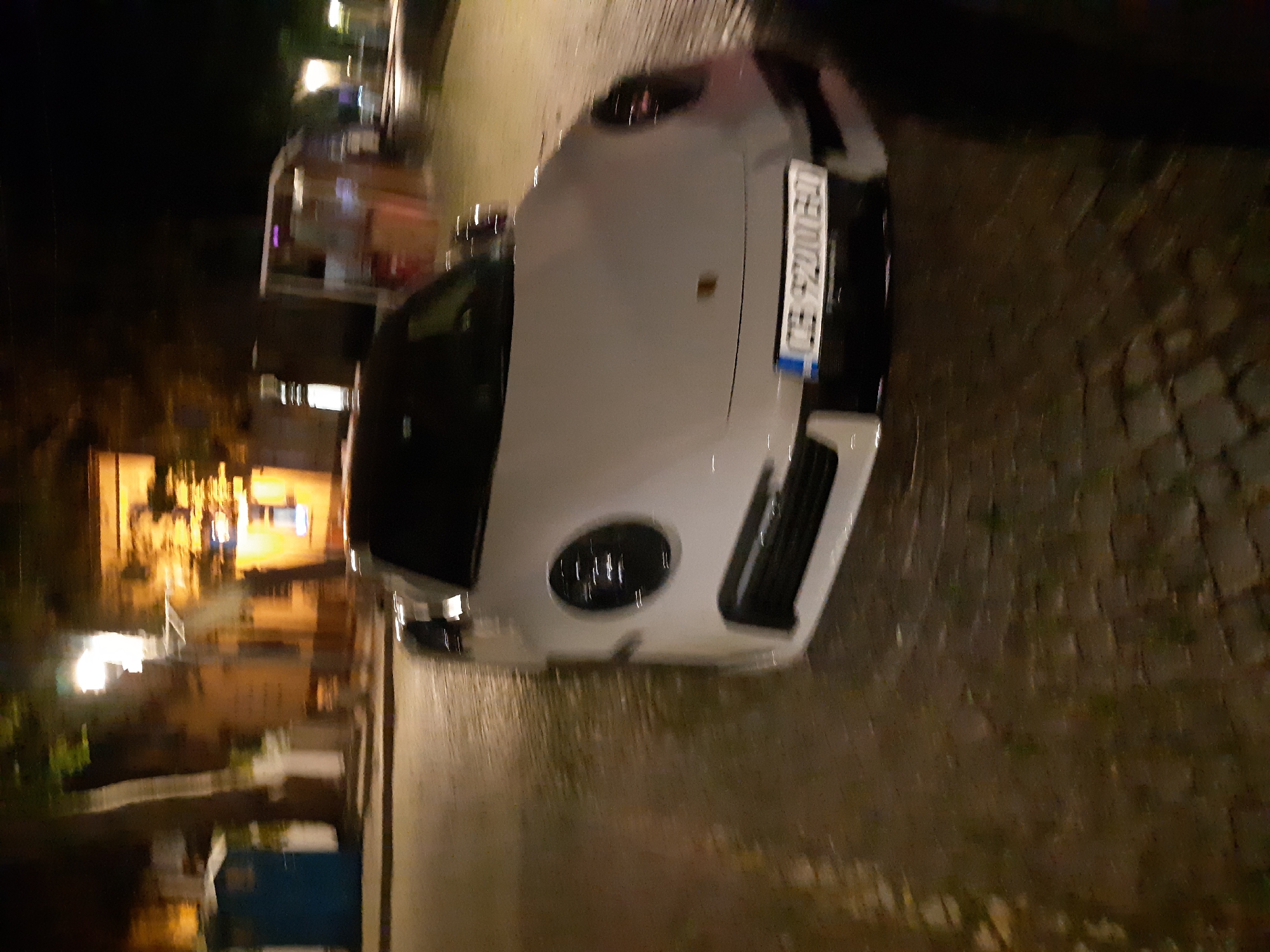 At night in Plovdiv, Bulgaria, a light-colored Porsche is parked on a cobblestone street. The vehicle faces the viewer. The license plate is partially visible. Behind the car is a building facade illuminated by streetlights. The street surface appears wet. There are no people visible in the immediate foreground.