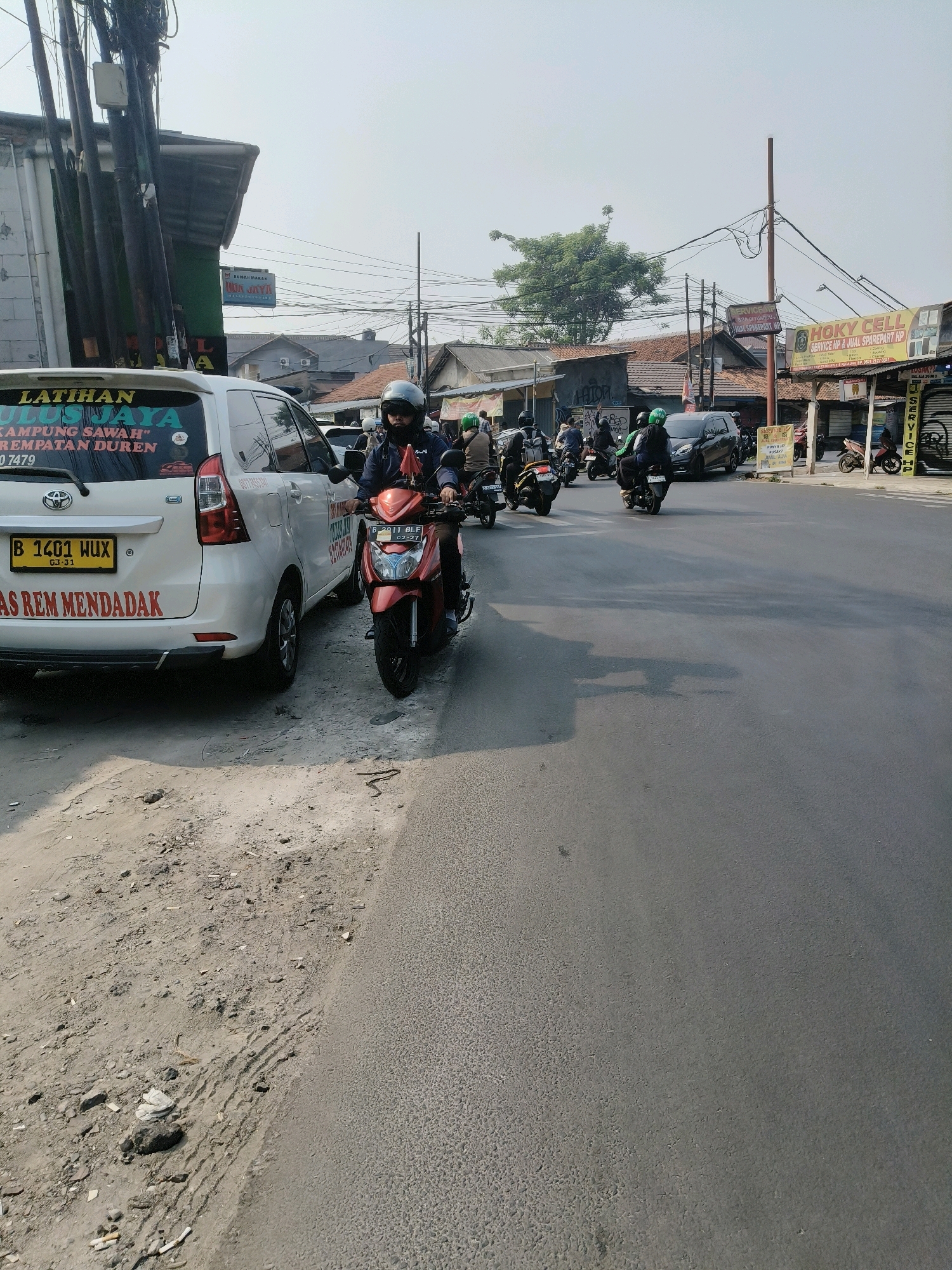 A street scene in South Tangerang, Indonesia, features a white Toyota Avanza parked on the left side of the road. The car has stickers on its rear window, including text that reads "LATIHAN ULUS JAYA" and "KAMPUNG SAWAH" with a phone number. A red motorcycle with a rider wearing a helmet is in the foreground, stopped on the road. The rider is looking forward. Several other motorcycles are visible on the street, with riders also wearing helmets and some with passengers. Shops and buildings line the right side of the street, including signs for "RUMAH MAKAN" and "HOKY CELL SERVICE HP & JUAL SPAREPART HP." The sky is overcast, and the road surface appears to be asphalt. The ground next to the car on the left is dusty and uneven, with scattered debris.