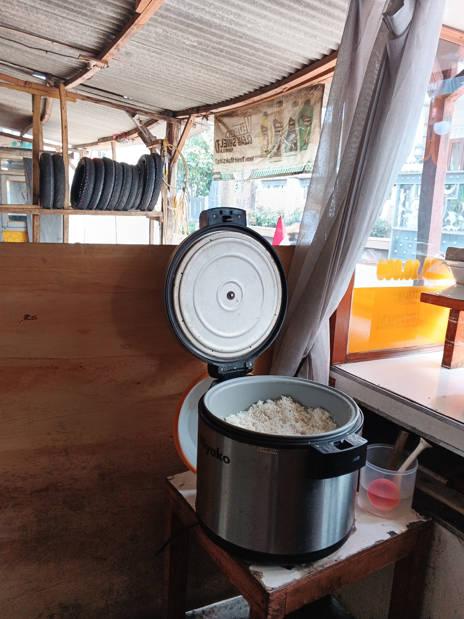 The image shows an interior scene, likely a food stall or small eatery in Gandasari, Indonesia. In the foreground, a silver rice cooker filled with cooked rice sits on a wooden table. The lid of the rice cooker is open, revealing a white inner pot. Next to the rice cooker is a clear plastic cup with a pink object inside, and a wooden stirring utensil. Behind the rice cooker, a wooden shelf holds a stack of black tires. The background features a corrugated metal roof and a large window with sheer curtains. A banner with text is visible on the wall behind the tires. The environment appears rustic and functional.