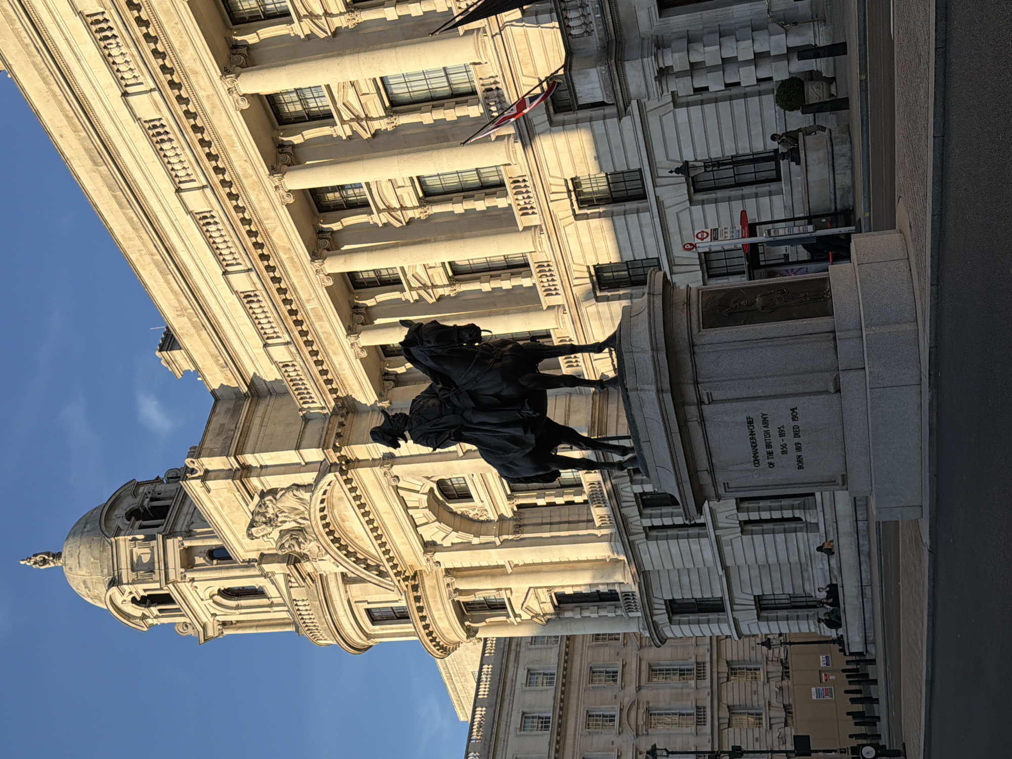 The image shows the Whitehall area of London, UK, featuring the statue of a commander-in-chief. The statue depicts a rider on a horse, standing on a stone plinth. In the background is a large, ornate building with a domed roof. Architectural details include columns, arches, and decorative embellishments. A Union Jack flag hangs from the building. A red London bus stop sign is visible next to the statue. Several people are standing near the statue's base and along the sidewalk in front of the building. The sky is blue.