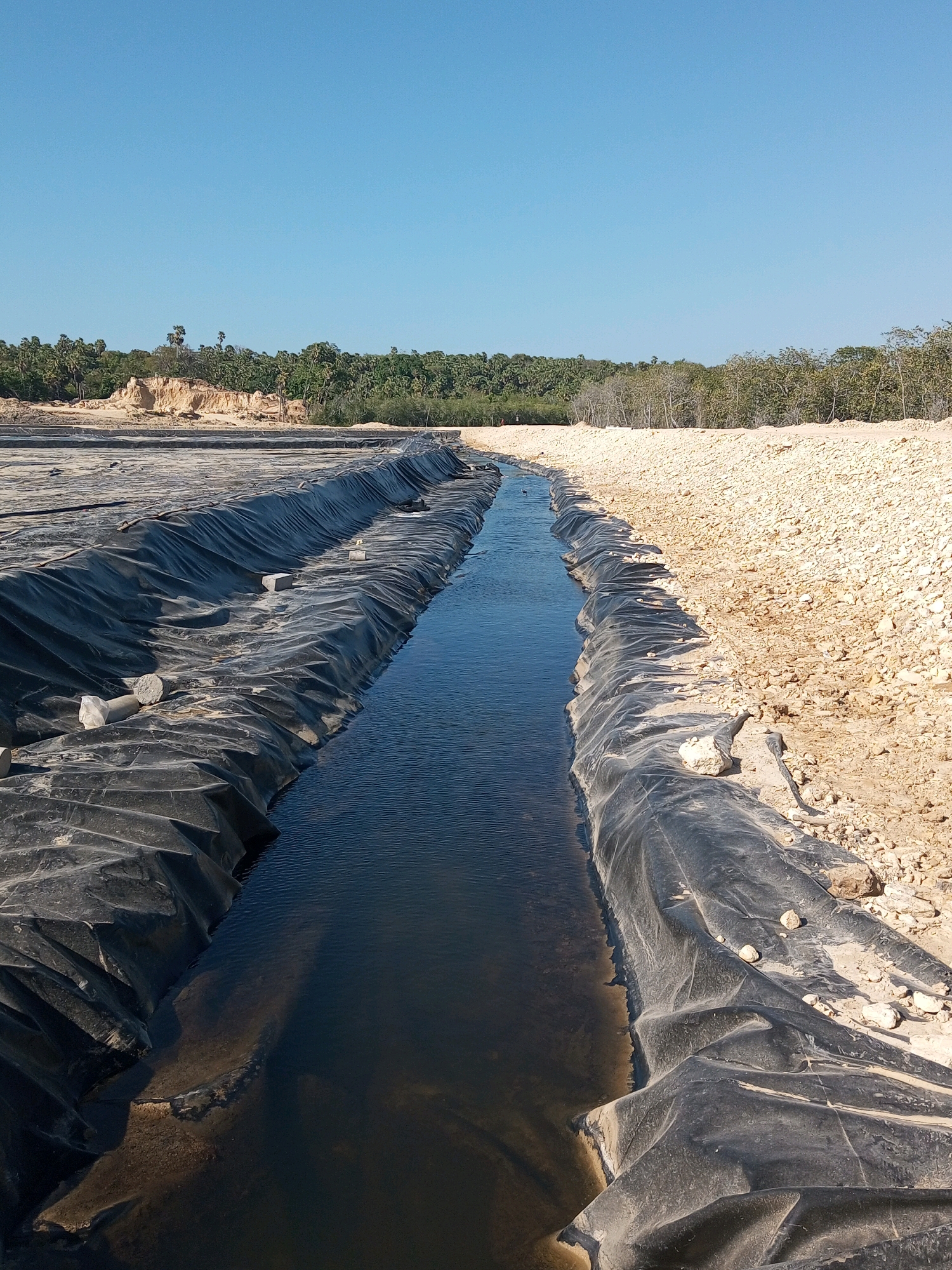 The image shows a construction site in an arid environment. A narrow channel of dark water flows through the center of the frame, lined by black plastic sheeting on both sides. The sheeting is wrinkled and appears to be a liner for a pond or canal. The land on the right is a mound of light-colored, coarse gravel and sand. The land on the left, further back, is a similar sandy embankment, with some rocks and debris visible. In the distance, a line of lush green trees and vegetation forms a stark contrast to the barren foreground. The sky is a clear, bright blue. The scene suggests a project involving water management or containment in a naturally dry area.
