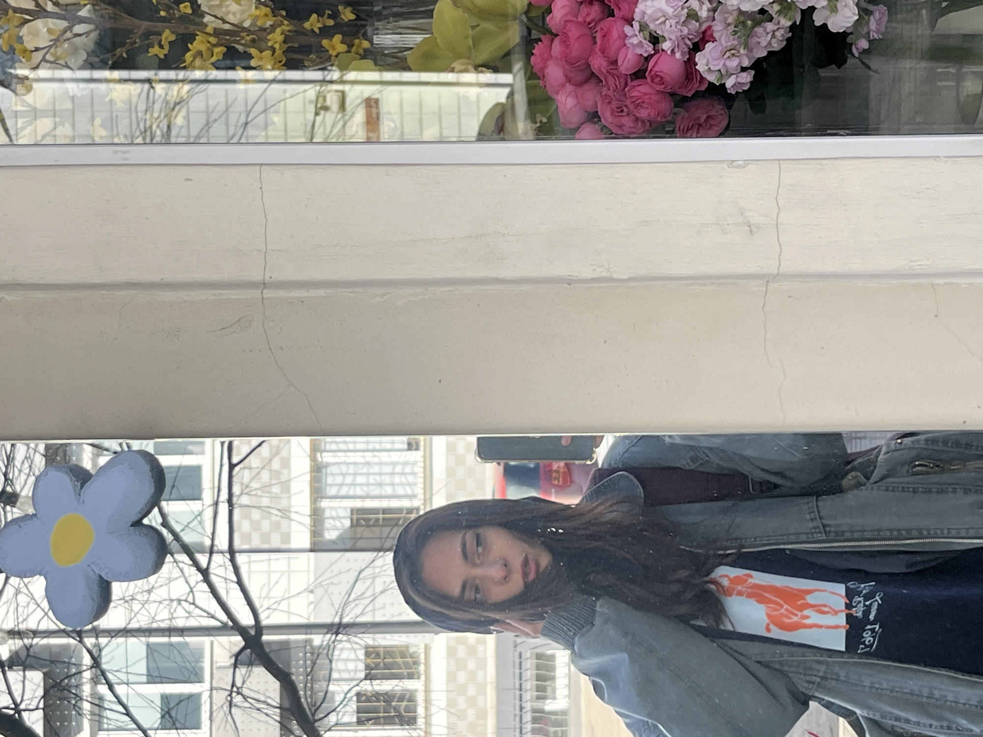 A reflection in a window captures a young woman wearing a blue jacket, a dark shirt with a prominent orange logo, and a burgundy top underneath. The reflection also shows an urban scene with buildings visible through bare tree branches and a large blue flower decoration in the lower left. Above, the window displays cut flowers and yellow flowering branches. Based on visual cues, it is plausible that the image might have been taken in Krasnoyarsk, Russia, given the architectural style of the buildings and the presence of bare trees, suggesting a late autumn or early spring season.