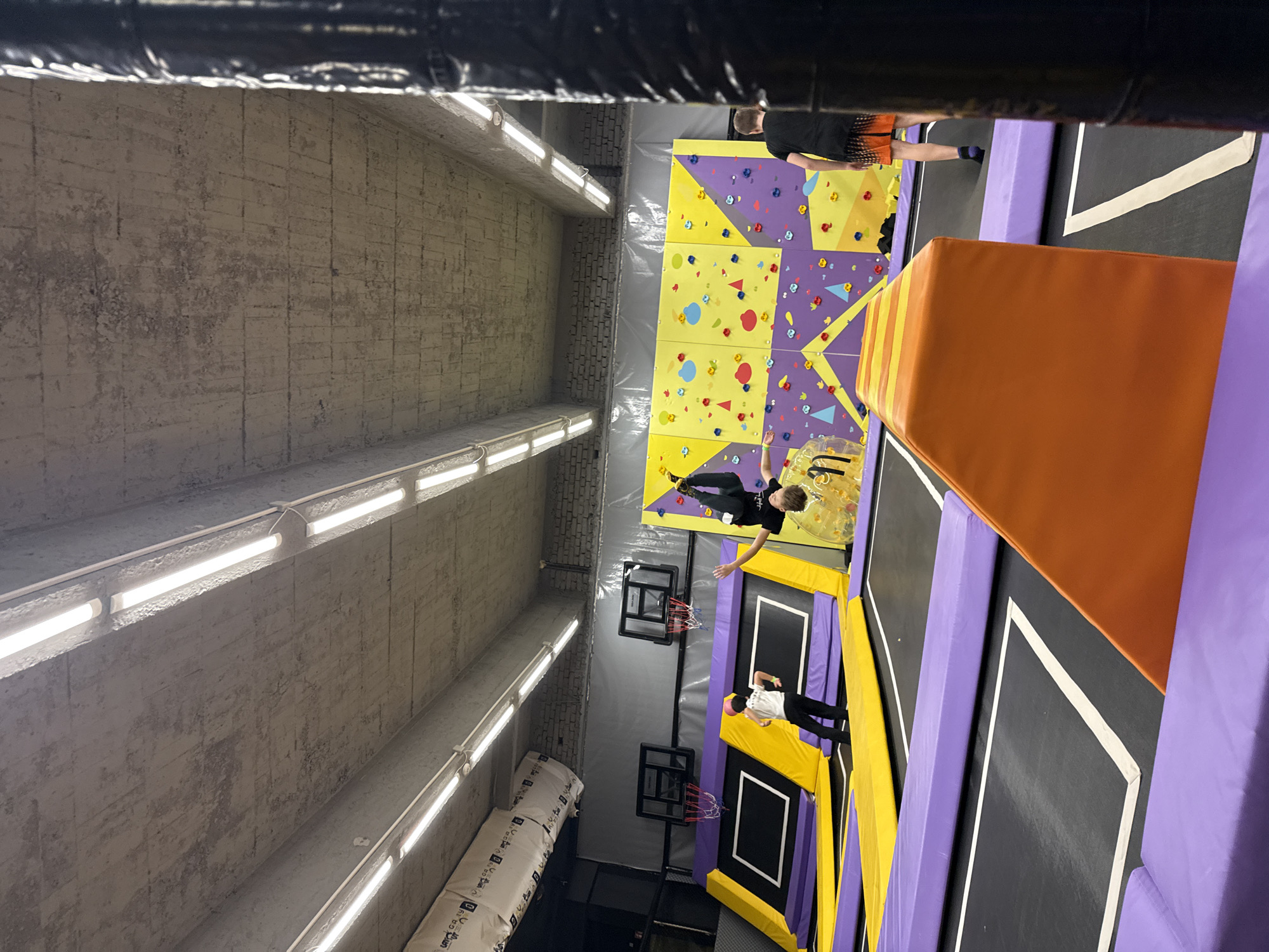 The image shows an indoor trampoline park in what appears to be Shumen, Bulgaria. The perspective is angled looking down, possibly from a balcony or elevated platform.
On the right, there is a large section of interconnected trampolines with purple and yellow safety pads. One person wearing a black shirt and shorts is standing on the edge of a trampoline near an orange safety pad.
Moving left, there are basketball hoops positioned above trampolines. A person wearing a white shirt is standing near a basketball hoop. Further along, there is a climbing wall with various colored holds. A person wearing a black shirt is reaching for the climbing wall.
The wall on the left is gray, exposed brick with rows of fluorescent lights running along its length. There is also a sign on the floor.
