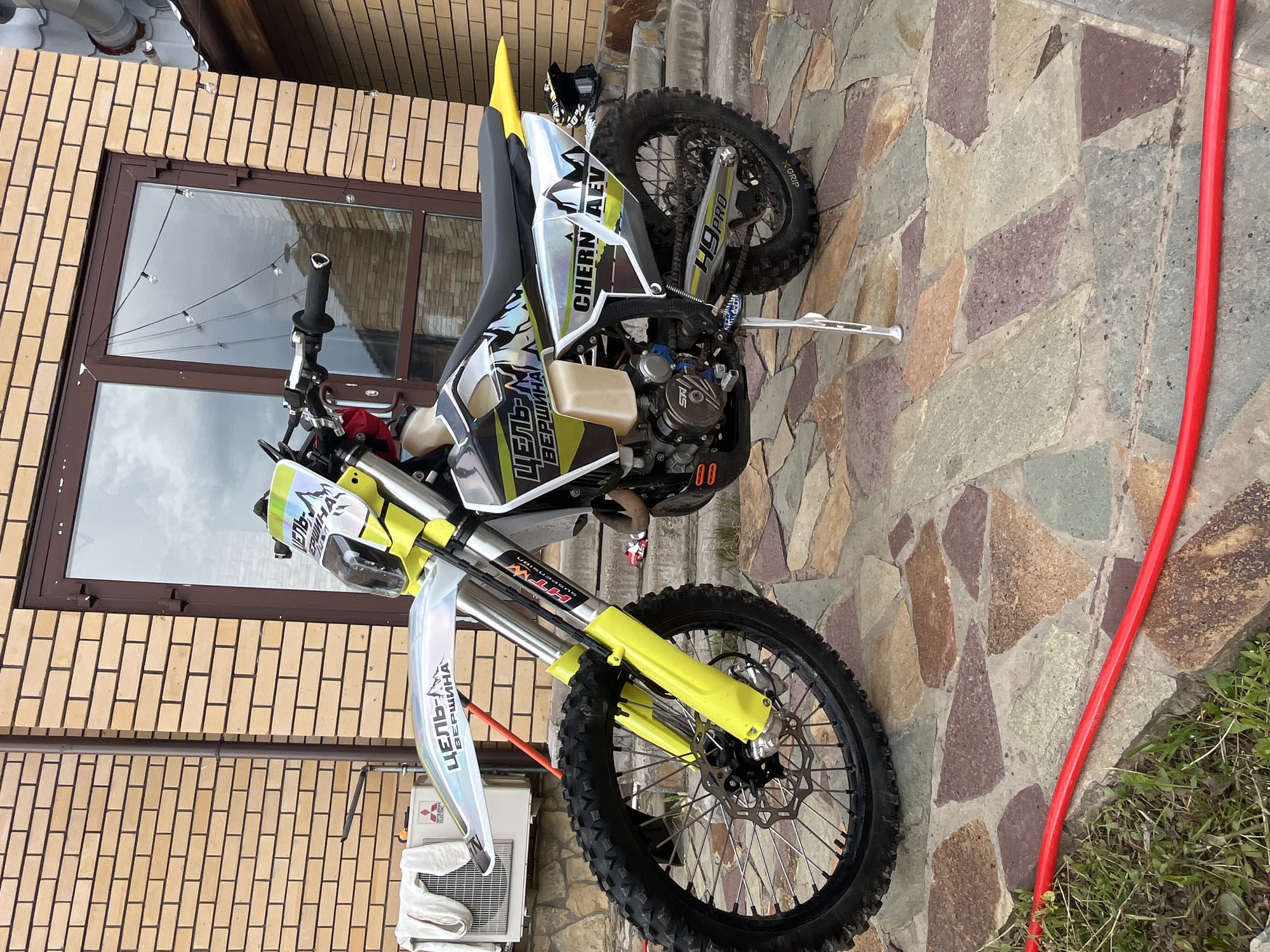 A yellow, black, and silver dirt bike or enduro motorcycle is parked on a stone-paved area next to a building in Ореховка, Russia. The motorcycle is presented in a landscape orientation, making it appear as if it's standing upright, resting on a silver kickstand. It features intricate graphics and decals across its body. A black helmet with white patterns rests on the rear seat.
The building behind the motorcycle has a lower section clad in irregular, multicolored stone tiles and an upper section made of light brown bricks. A large brown-framed window is visible, reflecting a cloudy or overcast sky, suggesting it is daytime. String lights are strung across the window frame. To the left of the motorcycle's front wheel, an outdoor air conditioning unit, likely a Mitsubishi, is mounted on the brick wall.
The ground surface consists of various shades of stone paving. To the right, a bright red hose snakes along the edge of the paved area, partially obscured by some green grass.
Visible text on the motorcycle includes "ЦЕЛЬ-М ВЕРШИНА" (Ziel-M Vershina), "HTW SUSPENSION" on the front forks, "CHERN AEV," and "H9PRO" on the rear swingarm. The motorcycle appears clean and well-maintained.