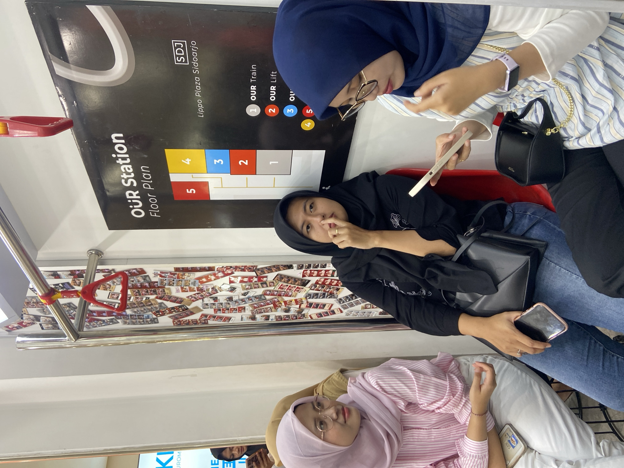 Three young women are captured in an indoor setting, likely a train or subway in Sidoarjo, Indonesia. The woman in the lower part of the image wears a light pink hijab and striped shirt, lying back on a seat and holding a phone. In the middle, another woman is dressed in black with a dark hijab, holding a phone in one hand and gesturing with the other. In the upper part of the picture, the third woman, wearing a blue hijab and glasses, is looking at a phone and wearing a light blue striped shirt. They are all holding their phones. In the background, there are numerous small photos pasted to the wall, and there's a sign that says "OUR Station Floor Plan Lippo Plaza Sidoarjo." Also, a red handrail is attached to the ceiling. The environment suggests public transport, decorated with photos and informational signage.