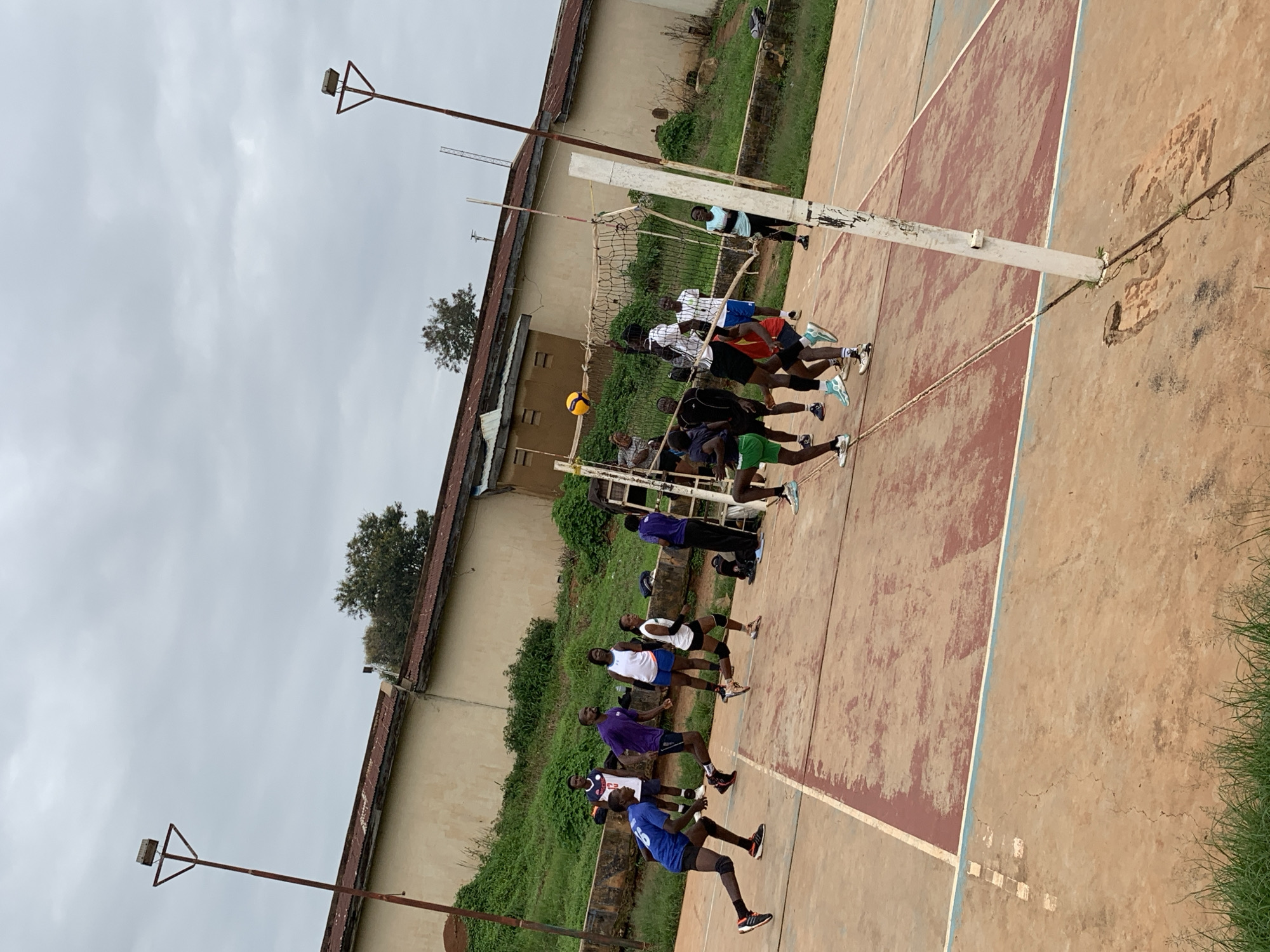 This is a medium-angle, eye-level shot of a group of people playing volleyball on an outdoor court. The court surface is made of a reddish-brown material with white and blue court lines. In the background, there is a building with a rust-colored roof and a light-colored wall. Green grass and trees are visible behind the building and around the court. Two tall, metal light poles with lights on top are present on the left and right sides of the frame.
There are approximately ten individuals actively engaged in the volleyball game. Most are wearing athletic attire, including shorts and t-shirts or tank tops. Their actions indicate they are mid-game, with some running, jumping, and reaching for the ball. A volleyball is visible in the air near the net, suggesting a serve or a spike is in progress. The overall atmosphere appears to be one of energetic sports activity. The location is identified as Gura Top, Nigeria.
