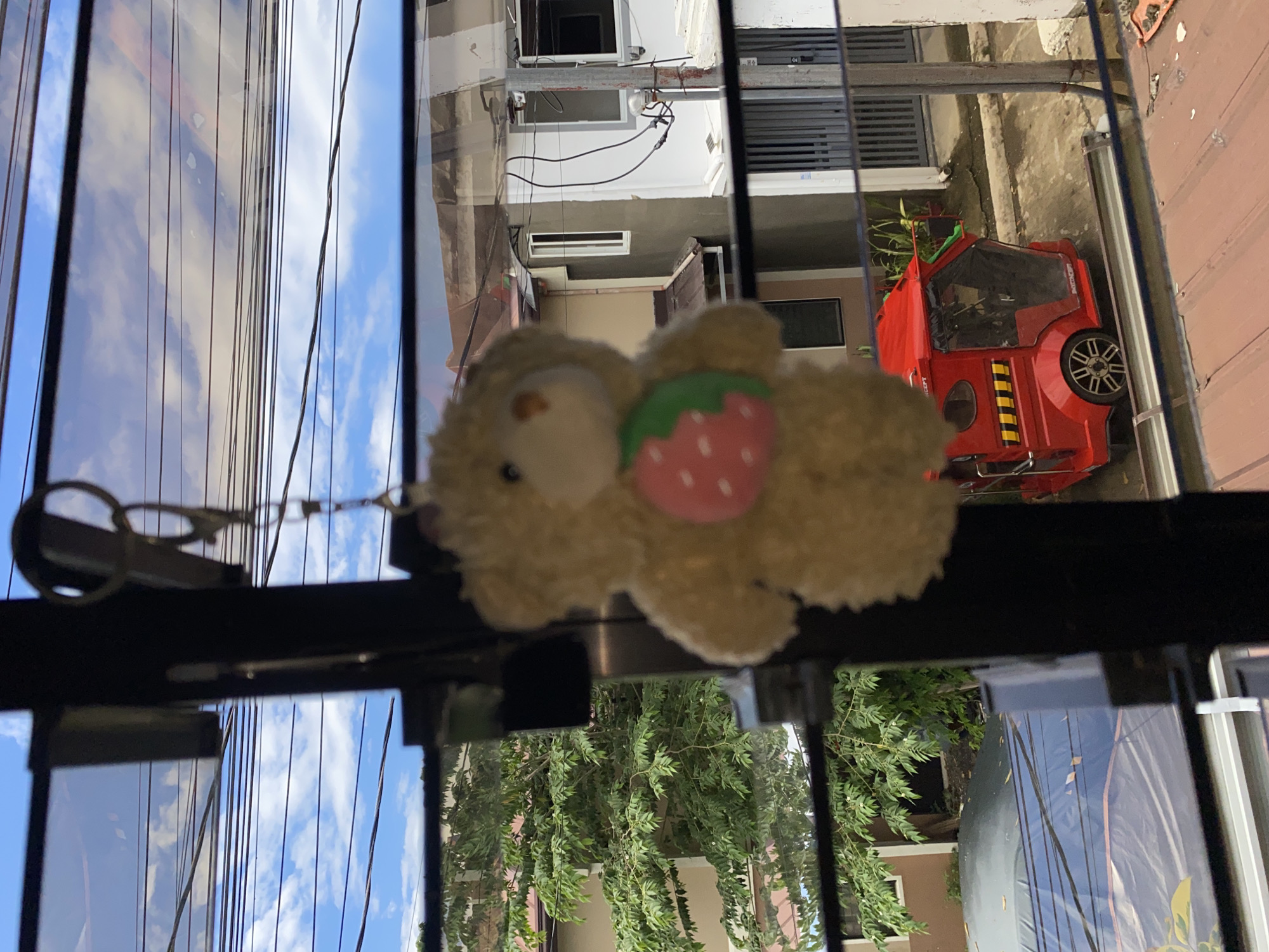A close-up, slightly blurry image shows a cream-colored teddy bear keychain with a pink strawberry applique hanging from a black frame, likely a window. The teddy bear is the primary subject, positioned in the foreground. Through the window, a scene of an urban environment in Davao City, Philippines is visible. A red, three-wheeled vehicle, commonly known as a "tricycle," is parked on a street. In the background, residential buildings and lush green trees are present, under a partly cloudy blue sky. The scene suggests an ordinary day in a residential area of the city.