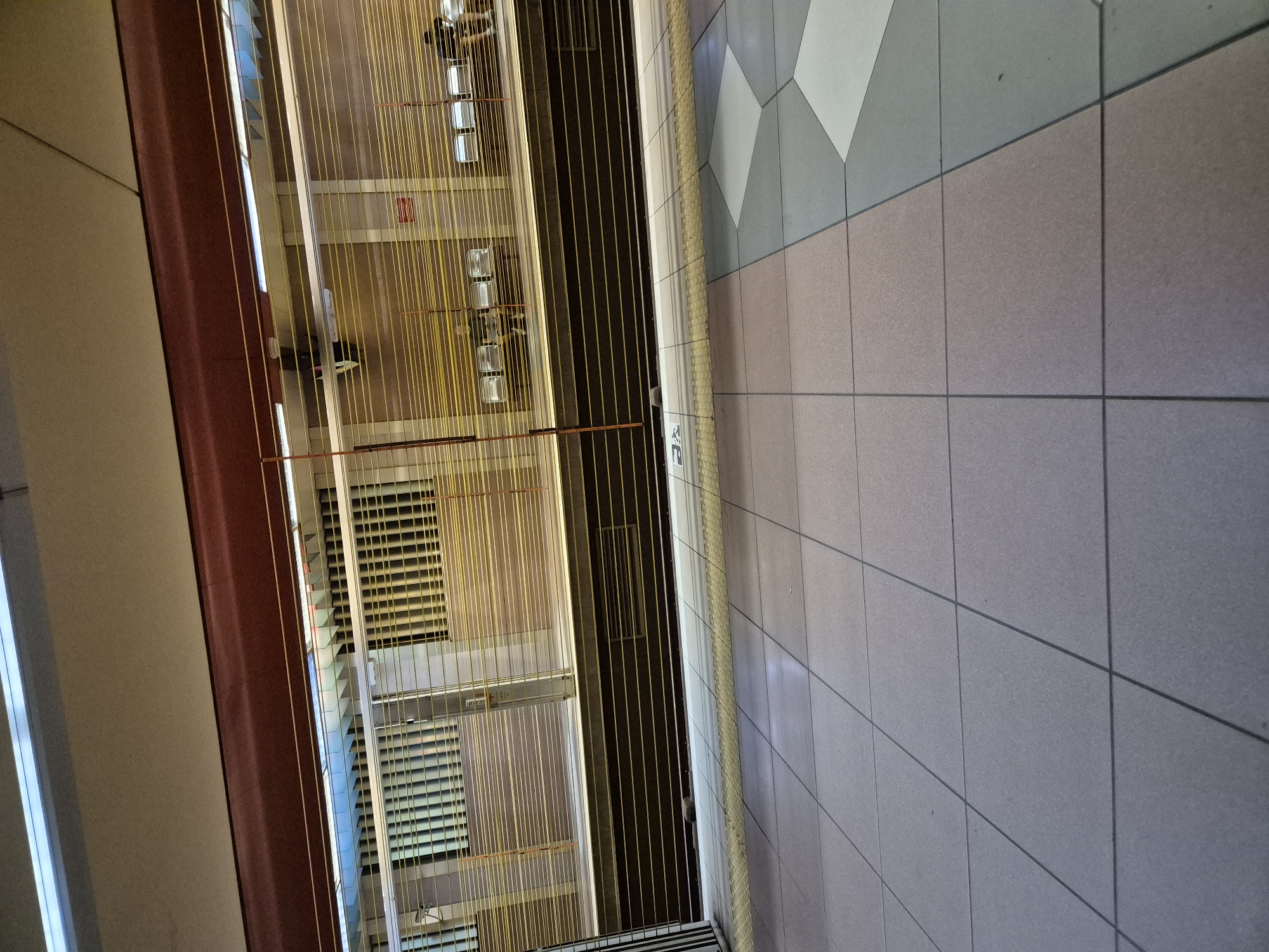 The image depicts a subway platform at the Sofia Central Railway Station in Sofia, Bulgaria. The platform is empty, with closed metal shutters obscuring the view of the tracks and the waiting area. The floor is tiled with a geometric pattern of light gray and pinkish-gray squares, with a yellow tactile paving strip along the edge of the platform. The ceiling is a pale beige, with linear fluorescent lights visible in the background. The overall impression is one of quiet and desolation, with the closed shutters suggesting that the station is not in operation.
