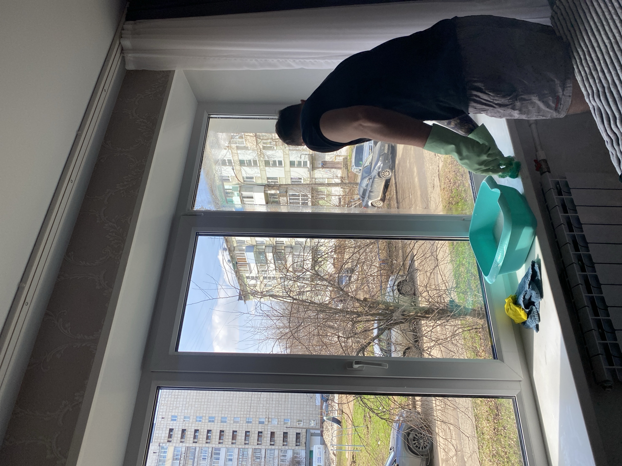 A person wearing gloves and a black t-shirt is cleaning a window. A light blue basin filled with water and a rag is on the windowsill. Outside the window is a view of apartment buildings and a car. The location is Chaikovsky, Russia.