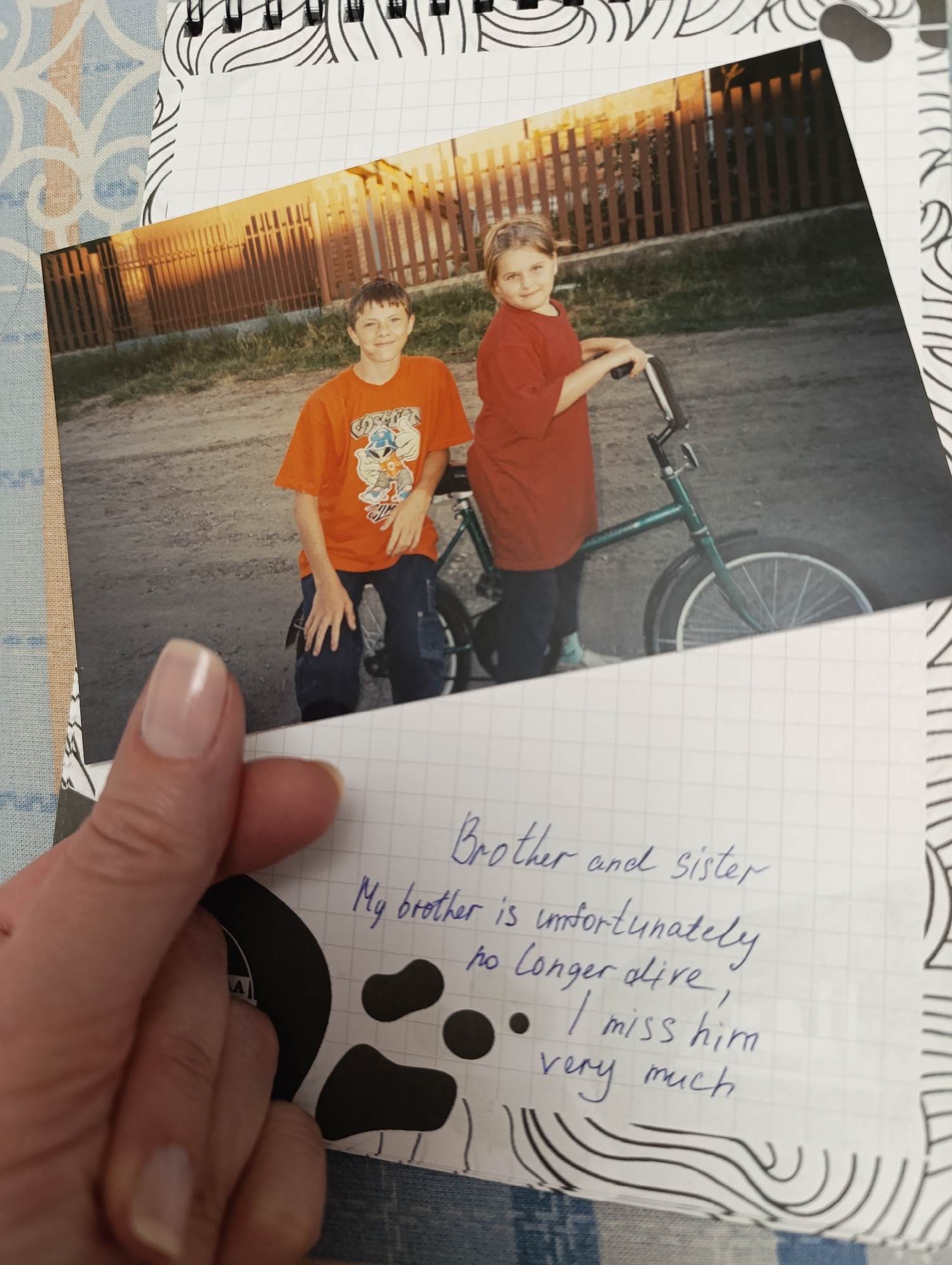 The image is a photograph of a brother and sister, likely taken in a rural or suburban environment. The photo shows the brother sitting on a bicycle and the sister standing next to him, holding the handlebars. The background consists of a dirt road, green vegetation, and a wooden fence. The photograph is placed on a sheet of lined paper with the handwritten text: "Brother and sister. My brother is unfortunately no longer alive, I miss him very much." A hand is holding the paper and the photo. The background includes a patterned surface and a notebook with a spiral binding.