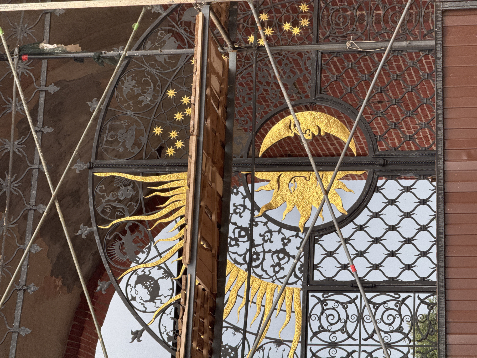 The image shows a detailed view of an ornate wrought-iron gate, possibly part of an entrance to a building or a historical landmark in Kazan, Russia. The gate features large, golden depictions of the sun and a crescent moon with a human-like face. The sun's rays are sharp and pointed, while the moon has a gentle expression. Surrounding these central figures are intricate metalwork designs, including zodiac signs or astrological symbols within circular frames and smaller golden stars. The gate is set against a brick wall, which is visible in sections behind the metalwork. Scaffolding is present in the foreground, obscuring parts of the gate and suggesting ongoing construction or restoration work. The overall impression is one of rich craftsmanship and symbolic artistry.