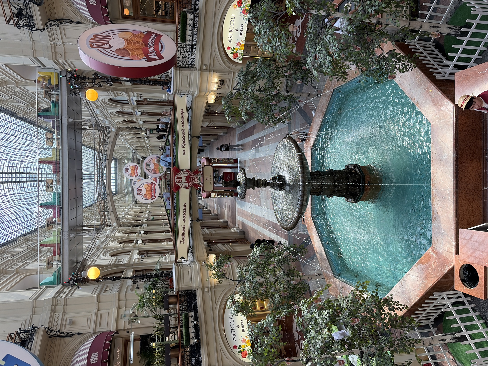 The image depicts the interior of the GUM department store in Moscow, Russia, a historic shopping arcade. A multi-tiered fountain with turquoise water is the central focus, positioned in the foreground. The fountain is surrounded by a pinkish marble basin and a low white picket fence. Artificial trees with small hanging decorations are placed around the fountain.
In the mid-ground, the arcade stretches out, featuring multiple levels of shops with arched entrances and balconies. Signs for various brands, including "ARTICOLI BOSCO" and what appears to be an ice cream shop with Cyrillic text "МОРОЖЕНОЕ," are visible. People are seen walking through the arcade and on the upper levels.
The upper part of the image reveals the distinctive glass-vaulted ceiling of GUM, allowing natural light to illuminate the interior. The architecture is ornate, with cream-colored facades and decorative railings. On one of the upper walkways, several people are seated at tables, possibly at a cafe.
In the lower right corner, a person wearing a straw hat and a dark red shirt is partially visible, looking towards the fountain. The overall scene conveys a bustling yet elegant atmosphere within the iconic Moscow landmark.