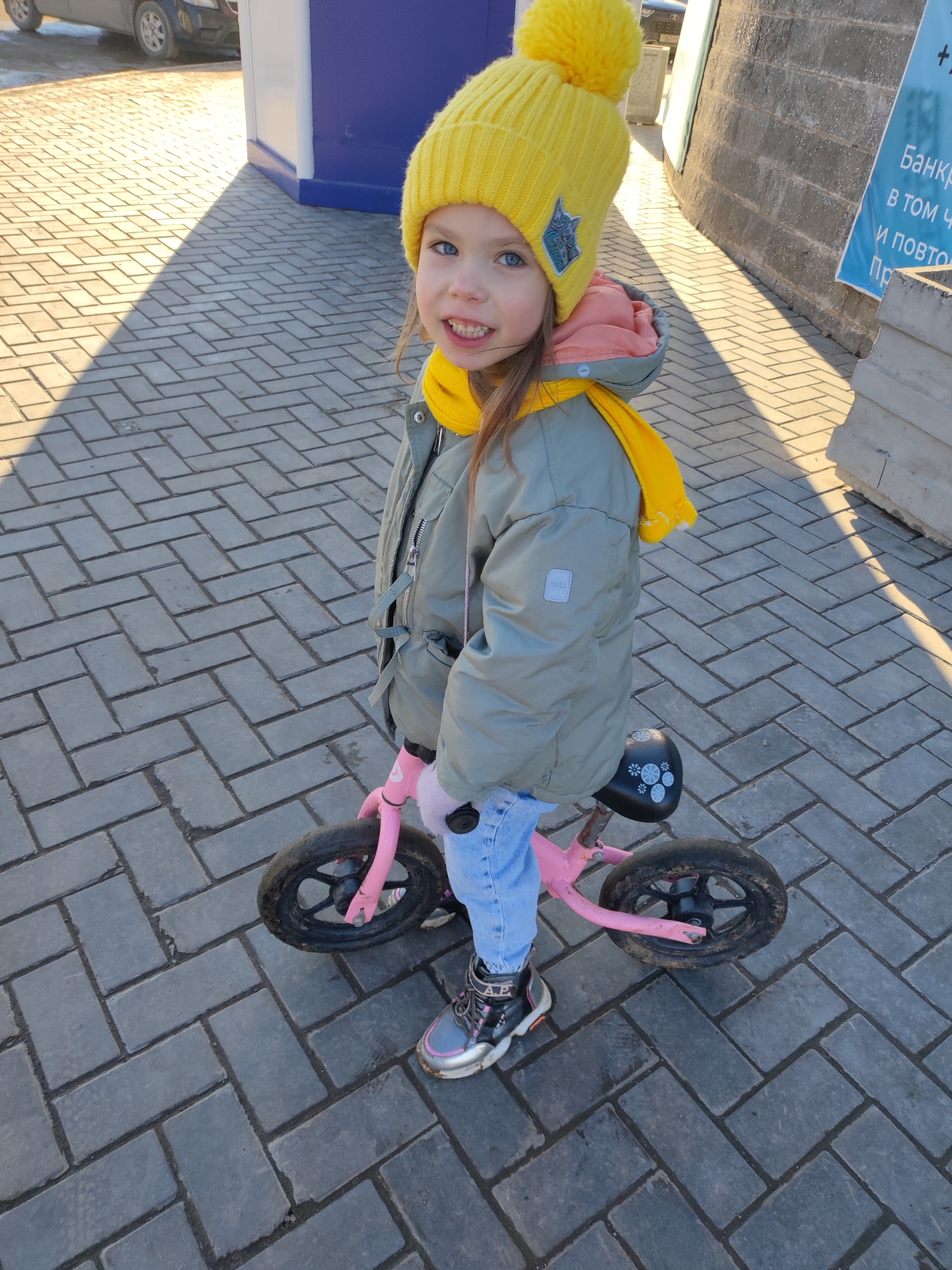 A young girl with blue eyes and a yellow hat stands next to a pink balance bicycle on a paved walkway. She wears a gray jacket and a yellow scarf. The location appears to be an urban environment, possibly in Votkinsk, Russia, indicated by the presence of a sign with Cyrillic text. There are buildings and a car in the background.