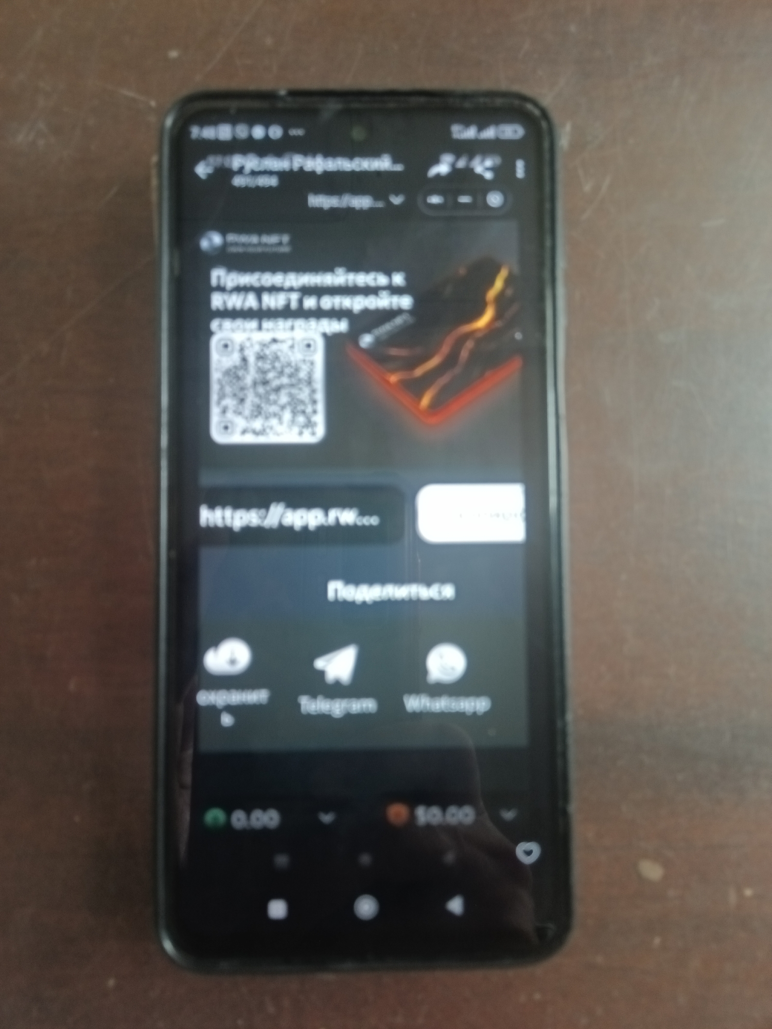 The image shows a smartphone held vertically on a wooden surface. The smartphone screen displays a webpage with Russian text and graphics related to NFTs. The top of the screen shows the time "7:48", battery and network indicators. The webpage includes a QR code, a web address "https://app.rw...", and icons for sharing via Telegram and WhatsApp. Below these, there are two currency indicators, "0.00" and "$0.00". The background of the image is a wooden table. There is no indication of the city of Kyiv or Ukraine in the image itself.