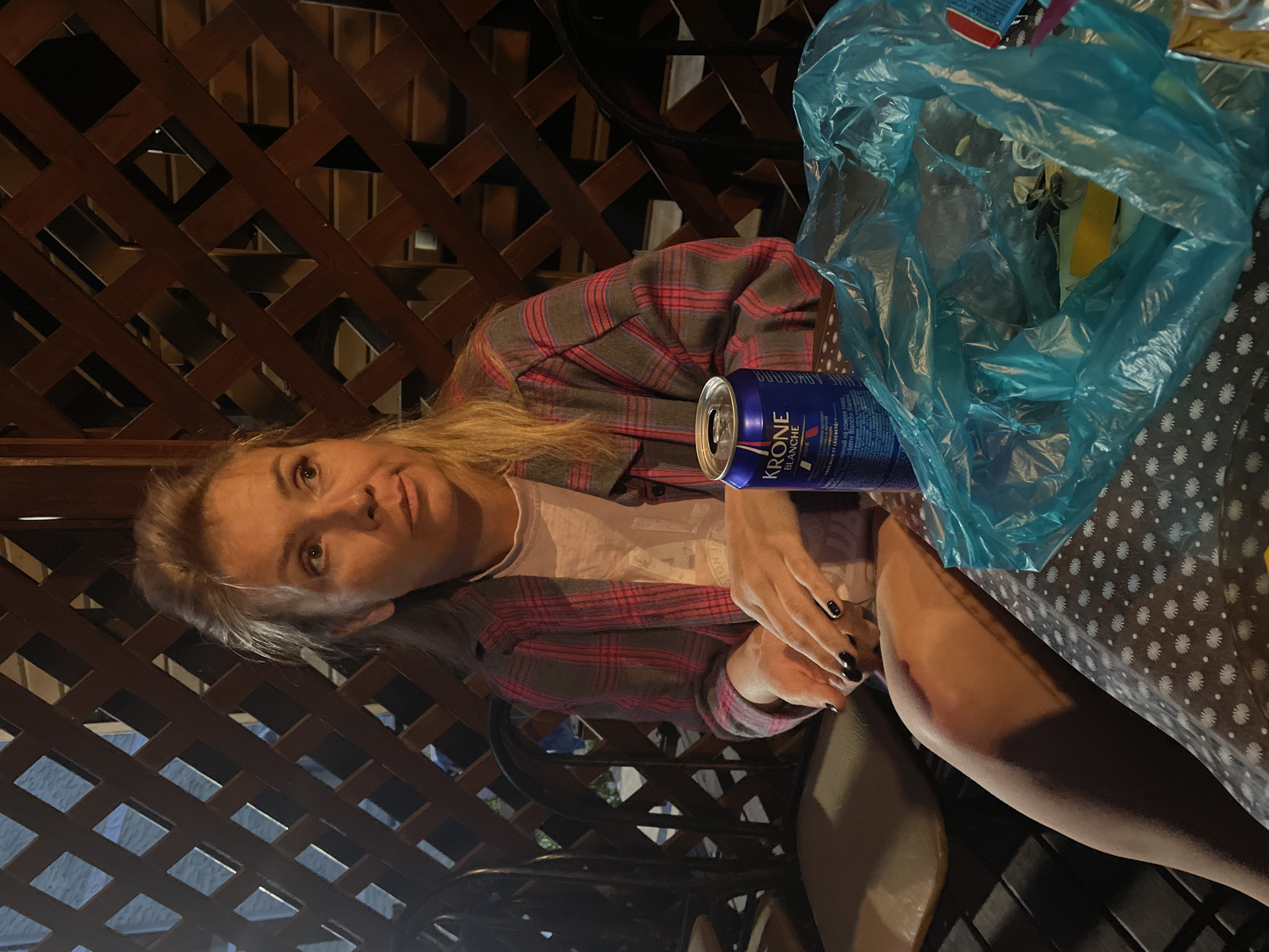 An outdoor shot shows a blonde woman sitting at a table on a patio. She is wearing a pink and grey plaid shirt over a white t-shirt. Her right hand is resting on the table, and her left hand is in her lap. A blue can of Krone beer is in front of her, and a bright blue plastic bag filled with various items sits to the right. The background features a wooden lattice fence and dark chairs. The lighting is dim, suggesting it is dusk or evening.