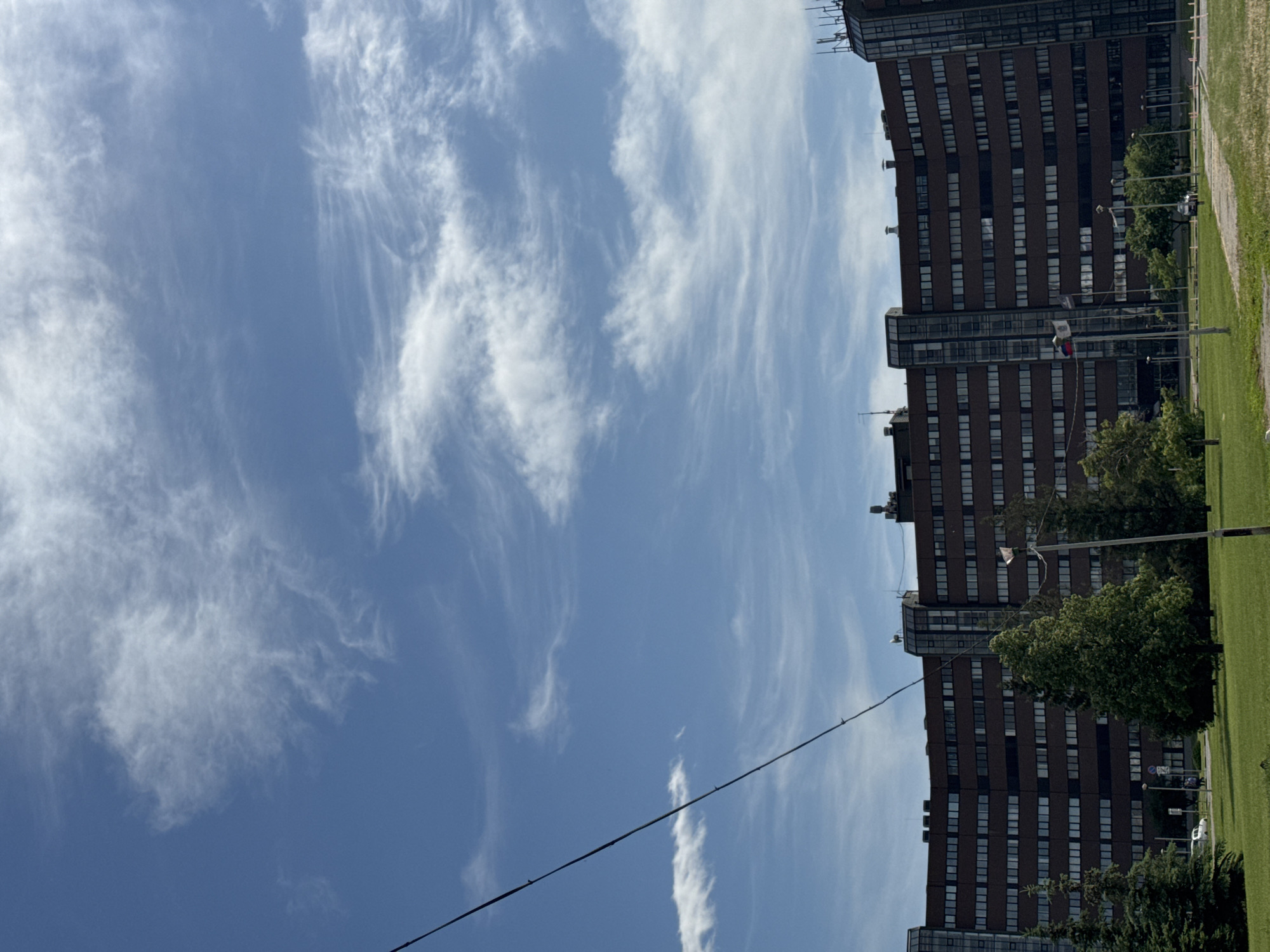 The image displays a daytime scene featuring a large, multi-story, dark brown building with a curved facade and numerous rectangular windows. Vertical glass architectural elements are visible at intervals. The building's roof features antennas and other structures. A bright blue sky with scattered white, wispy cirrus clouds occupies the upper portion of the frame. A dark cable or utility line traverses diagonally from the top left towards the bottom center. The foreground consists of a expansive green lawn with several trees, including both evergreen conifers and deciduous trees with full green foliage. A paved road or pathway is visible in the mid-ground, where a white car and a darker vehicle are present. A light-colored pole stands in the mid-right foreground, and another pole closer to the building displays two flags, one resembling the Czech national flag. Two indistinct figures, presumably people, are walking on a paved area in front of the building on the left side. The location is an urban or suburban setting in Unknown, Unknown.