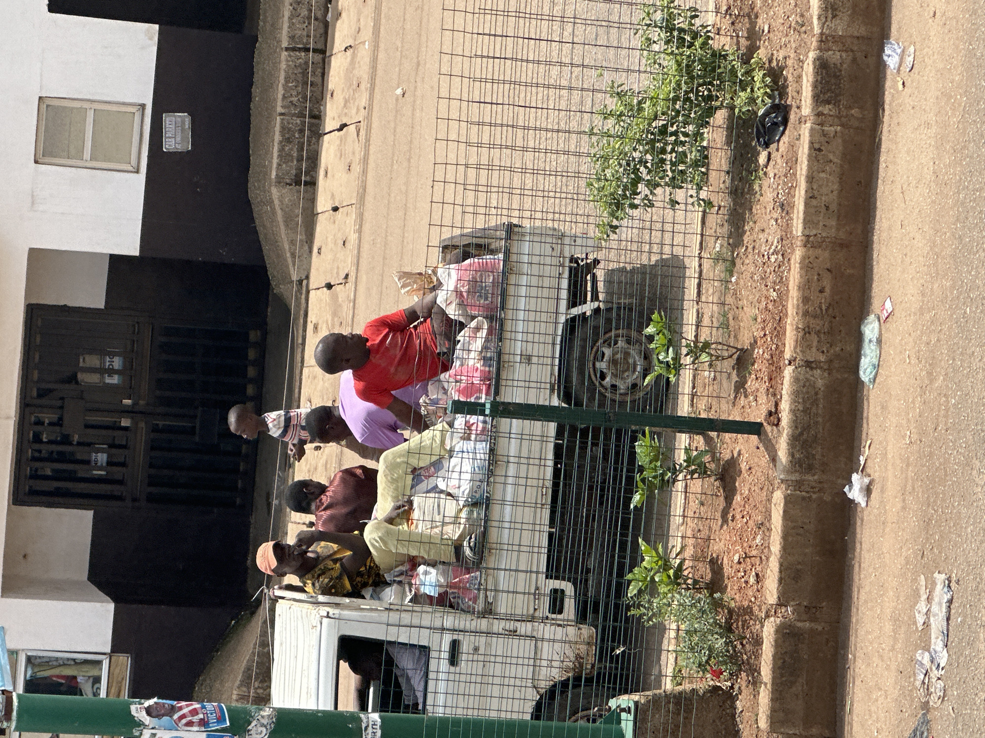 In Bukuru, Nigeria, a white pickup truck is parked on a dirt road. The truck bed is filled with various bags and items. Five individuals are visible in and around the truck. A child in a striped shirt stands to the right of the truck. Three men are seated in the truck bed, one wearing an orange cap and a patterned shirt, another in a maroon shirt, and a third in a red shirt. Another person, partially obscured, is seated in the truck's cabin on the left. A metal fence runs along the foreground, separating the road from the truck. The background shows a building with a black and white facade and gated entrances. The area appears to be an urban environment with some sparse vegetation and scattered litter on the ground.