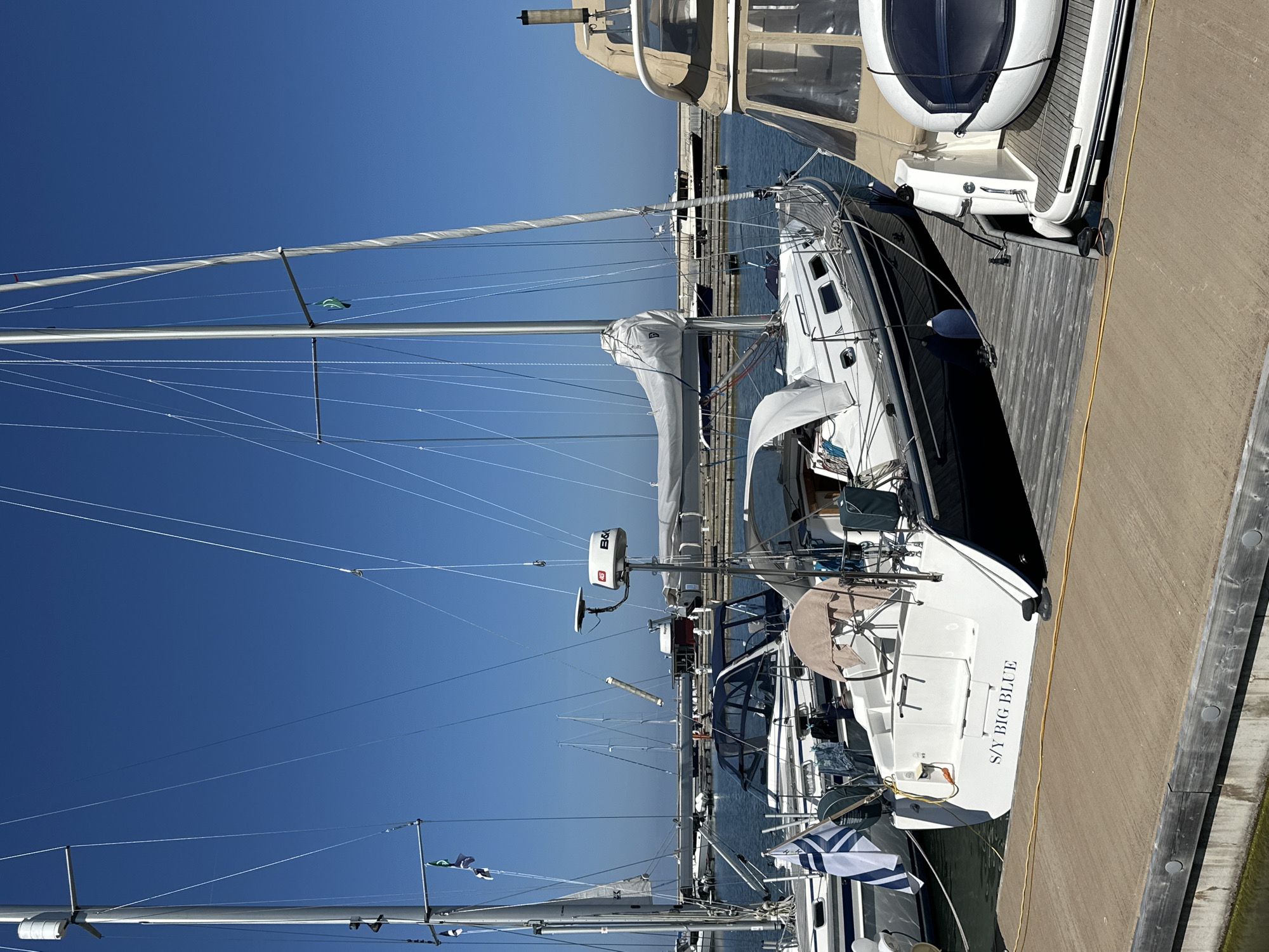 This is a photograph taken on a clear day in the city of Tallinn, Estonia, likely in the harbor or marina area. The foreground features a wooden dock and a portion of another boat. The main subject is a white sailboat named "S/Y BIG BLUE", docked alongside other vessels. The sailboat has a dark blue hull and a covered boom. Several other sailboats and a larger motor yacht are visible in the background, moored in the water. The sky is a vibrant, cloudless blue. The scene appears peaceful and typical of a marina on a sunny day.