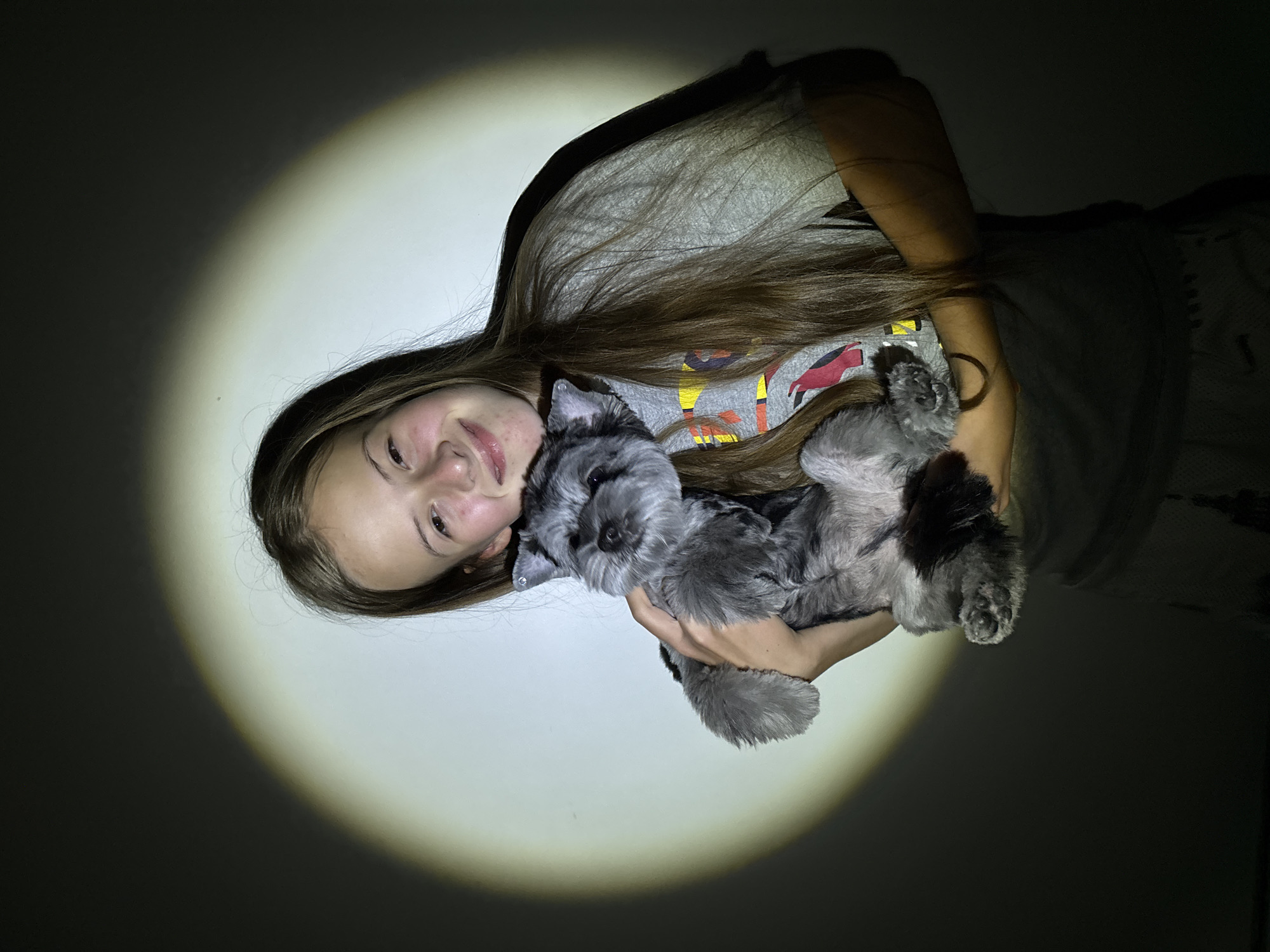 A young person, appearing to be a girl or young woman, with long, straight brown hair and a warm smile, is prominently featured in the image. She is holding a small, scruffy dog with grey and black fur, possibly a Schnauzer or terrier mix, cradled in her arms. Both subjects are looking towards the camera.
The young person is wearing a light-colored t-shirt with a partially visible graphic design featuring colorful shapes, including red and yellow elements. The dog has dark eyes and distinctive whiskers.
The scene is dominated by a large, bright, circular spotlight that illuminates the subjects, standing in stark contrast against a predominantly dark background. This strong lighting effect suggests the photo was taken at night or in a very dimly lit indoor environment, creating a focused and intimate portrait. The joyful expression on the person's face and the calm, attentive look of the dog convey an affectionate and happy moment.
Given the location context of Chaikovsky, Russia, the setting is likely an indoor space. There is no visible text in the image.