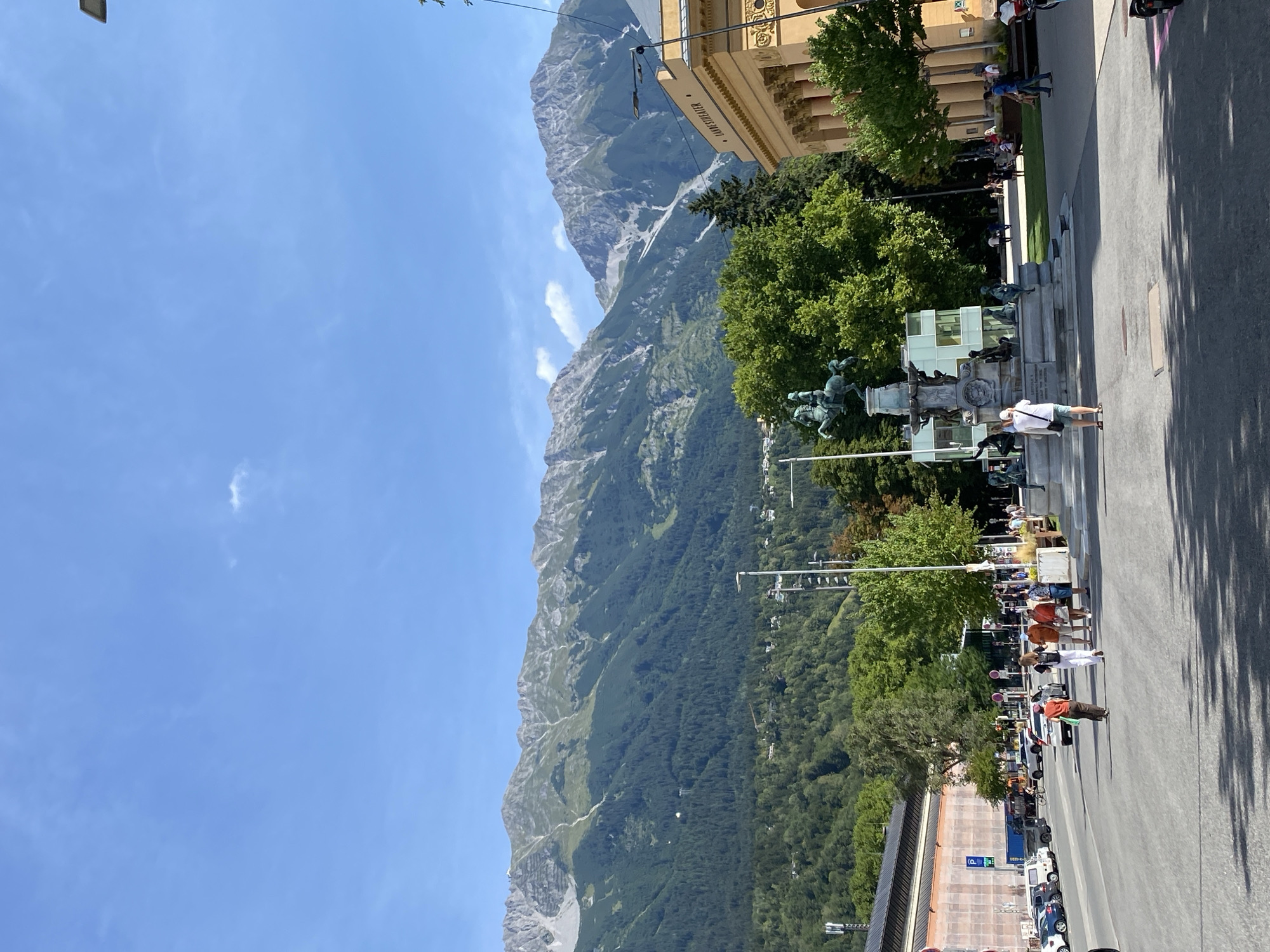 The image depicts a street scene in Innsbruck, Austria, with a prominent equestrian statue at a roundabout in the foreground. The statue is of a man on horseback, surrounded by other figures and architectural elements. Behind the statue, a grand building with classical columns and the inscription "LANDESKAPELLE" is visible. The street is paved, and there are cars parked along the side, as well as people walking on the sidewalks. In the background, lush green trees frame the scene, and beyond them, the majestic, rugged mountains of the Alps rise against a clear blue sky with a few scattered clouds. The overall impression is of a sunny day in a picturesque European city, blending urban life with the natural beauty of the surrounding landscape.