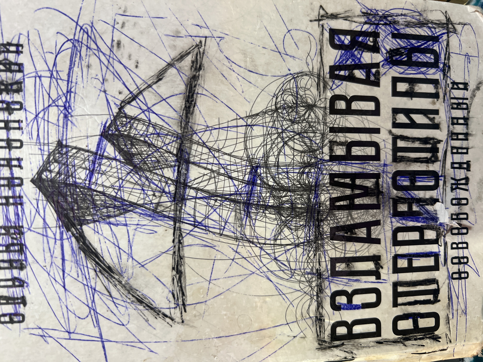 The image shows a close-up of a worn, possibly scratched, surface featuring abstract drawings and Russian text. The dominant drawing consists of dense black and blue scribbled lines forming geometric shapes. The black lines create a web-like structure resembling a partially defined object, possibly architectural or abstract. The blue lines appear more scattered, serving as background elements.
To the right and left of the drawing are two columns of black Russian text oriented vertically. The text on the left and right of the image is written in Cyrillic letters. The text appears bold and slightly condensed.
The surface of the image shows signs of aging and wear, including scratches, smudges, and discoloration. The lighting is somewhat uneven, casting subtle shadows across the surface.
There is not enough information in the image to determine its specific location in the city of Ильинская Поляна Снт, Russia.