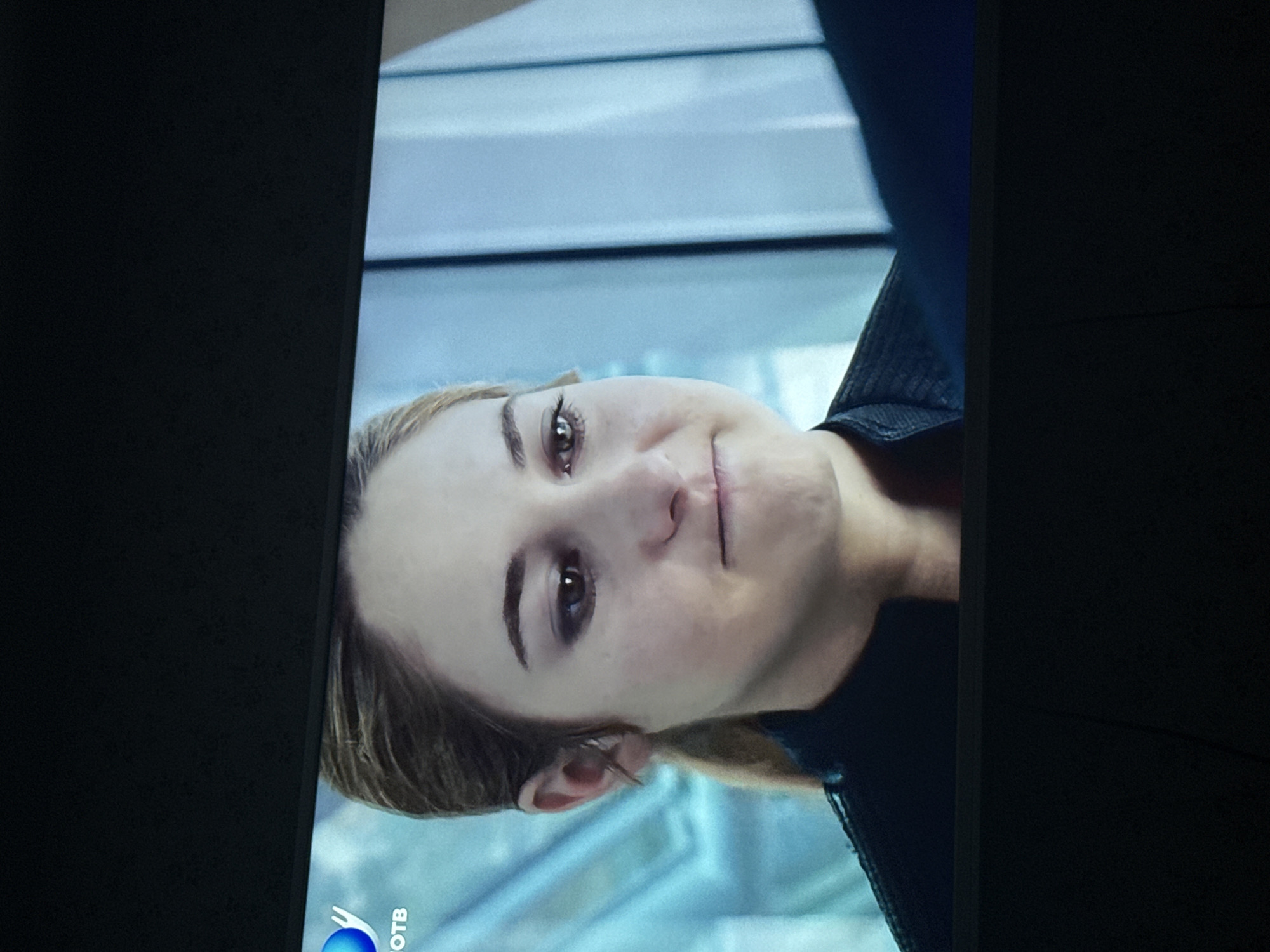 A medium, close-up shot shows a fair-skinned woman with brown eyes and blond hair. She is wearing a dark-colored shirt or jacket. The background is a light-blue color with a blurred design. A logo, possibly a television station, is visible in the lower-left corner with the letters "OTB" displayed. The image is slightly rotated counterclockwise. There is no information to establish the location as Mytishchi, Russia.