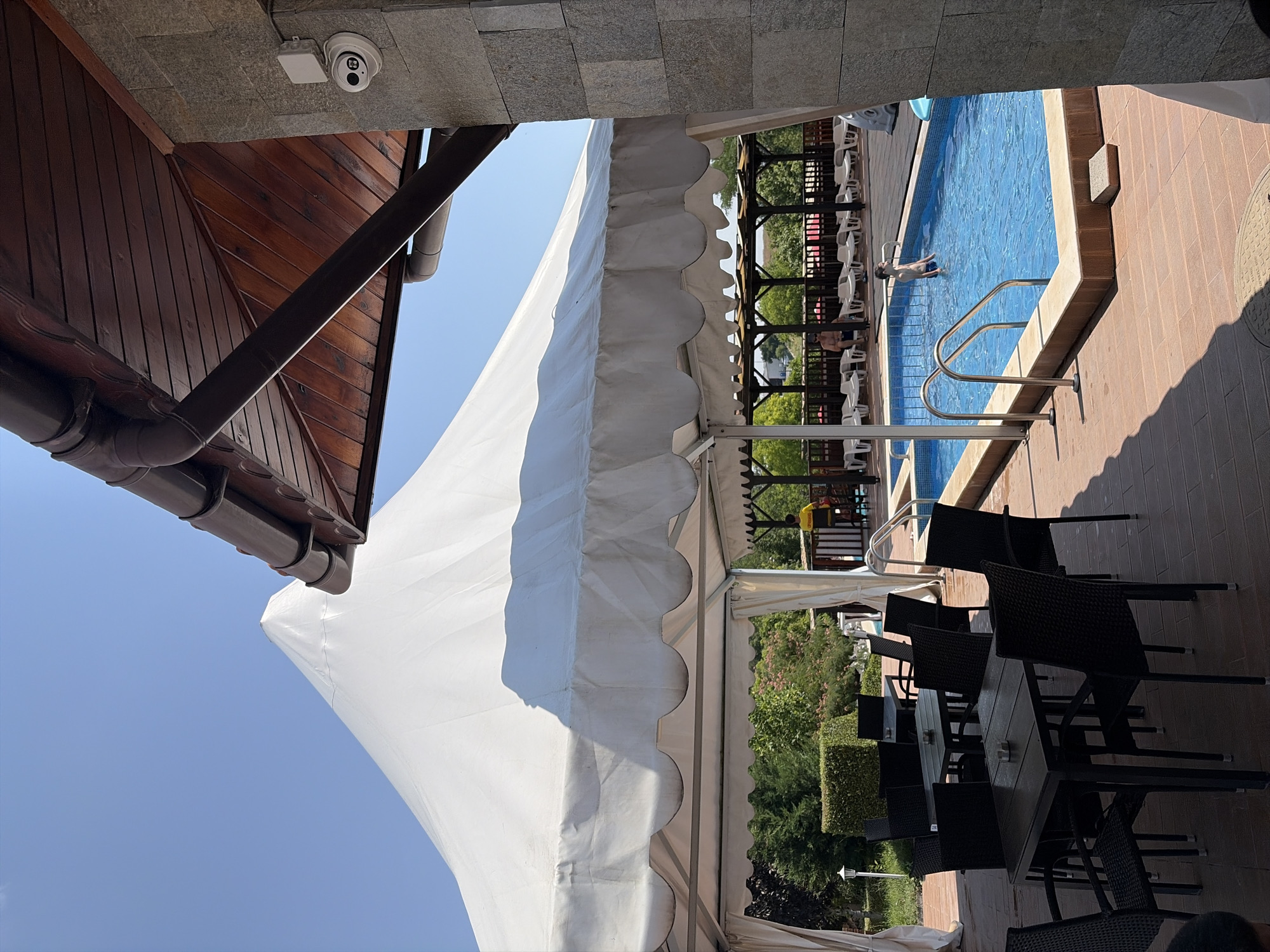 The image depicts an outdoor seating area adjacent to a swimming pool, likely located at a resort or hotel in Simeonovo, Bulgaria. A large white tent covers a section of the seating area, which features several black tables and chairs. In the background, a swimming pool is visible, with a young boy standing in the water. Lounge chairs are arranged around the pool, and lush greenery surrounds the area. The scene is bathed in sunlight under a clear blue sky.